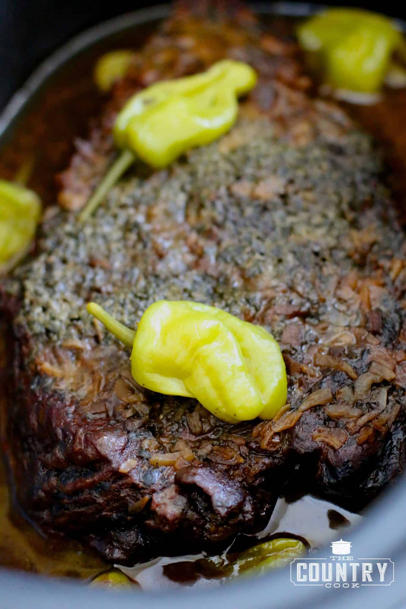 fully cooked whole pot roast in its juices in a slow cooker topped with peperoncini peppers.