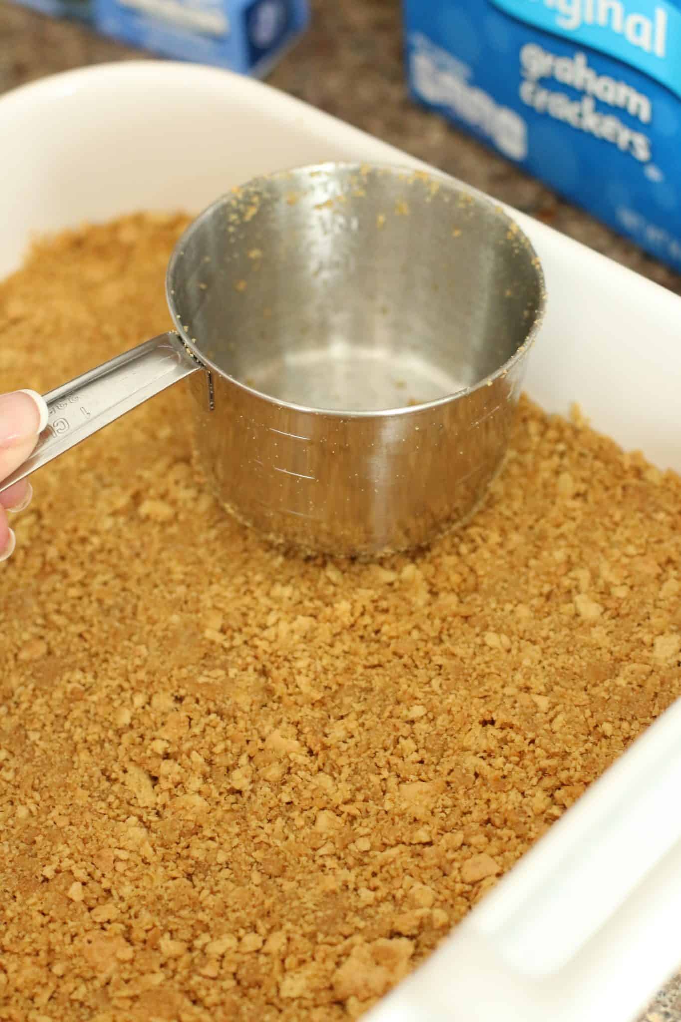 pressing graham cracker crumbs in cake pan with a measuring cup