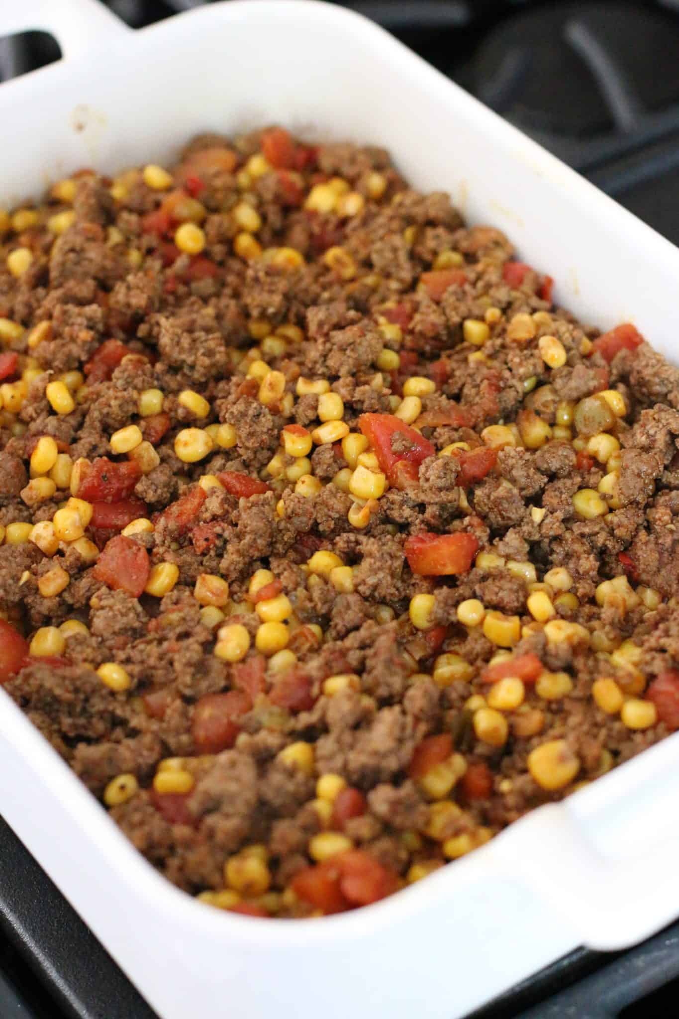 cooked ground beef mixture spread out on top of cornbread in a white baking dish.