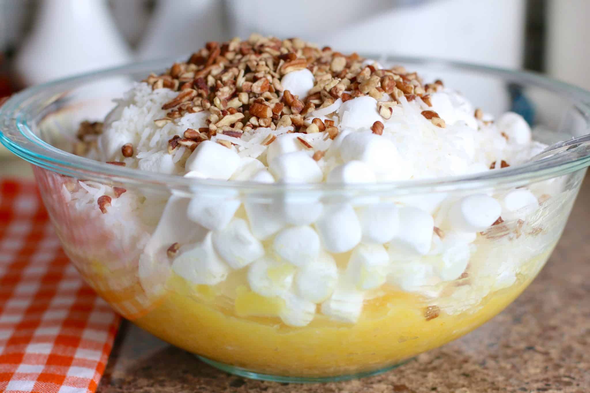 Cool whip, marshmallows, coconut and pecans added to pineapple mix in clear bowl.