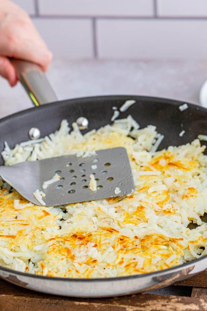 a spatula pushing down shredded hash browns in a large skillet. 