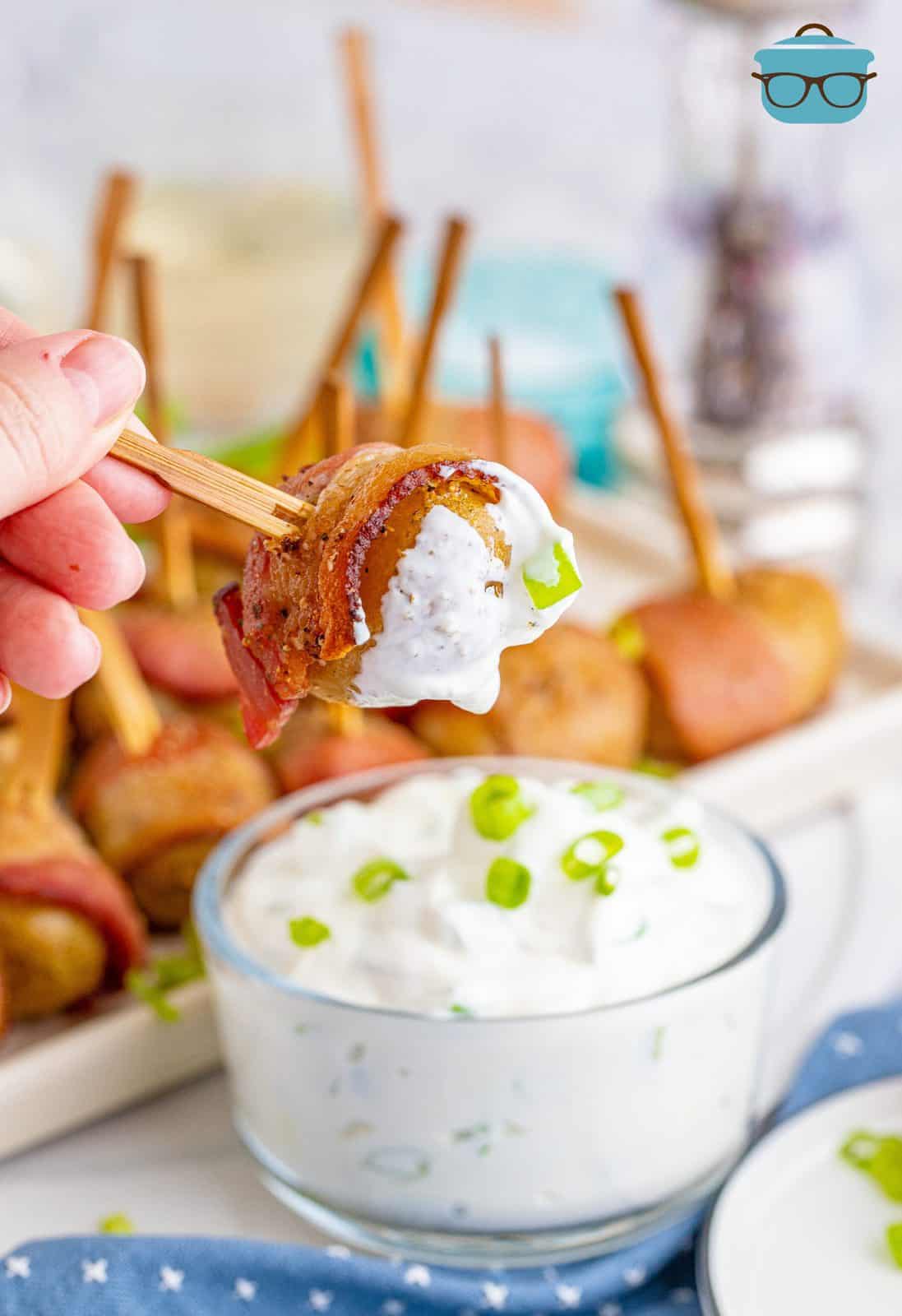 a hand dipping a bacon wrapped tomato into sour cream and green onion dip. 