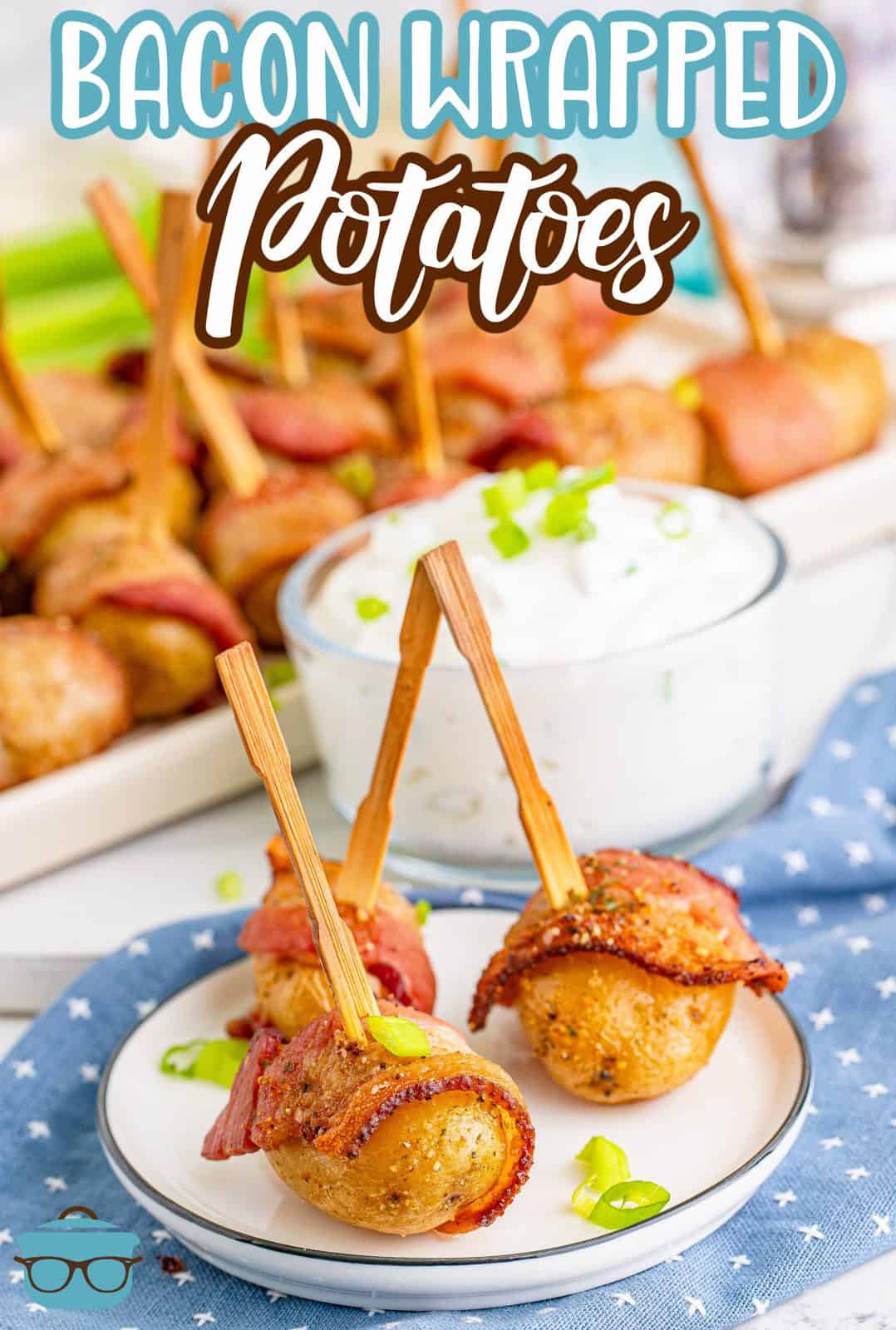 three bacon wrapped potatoes with toothpicks on a white plate in front of a tray holding several more bacon wrapped potatoes.