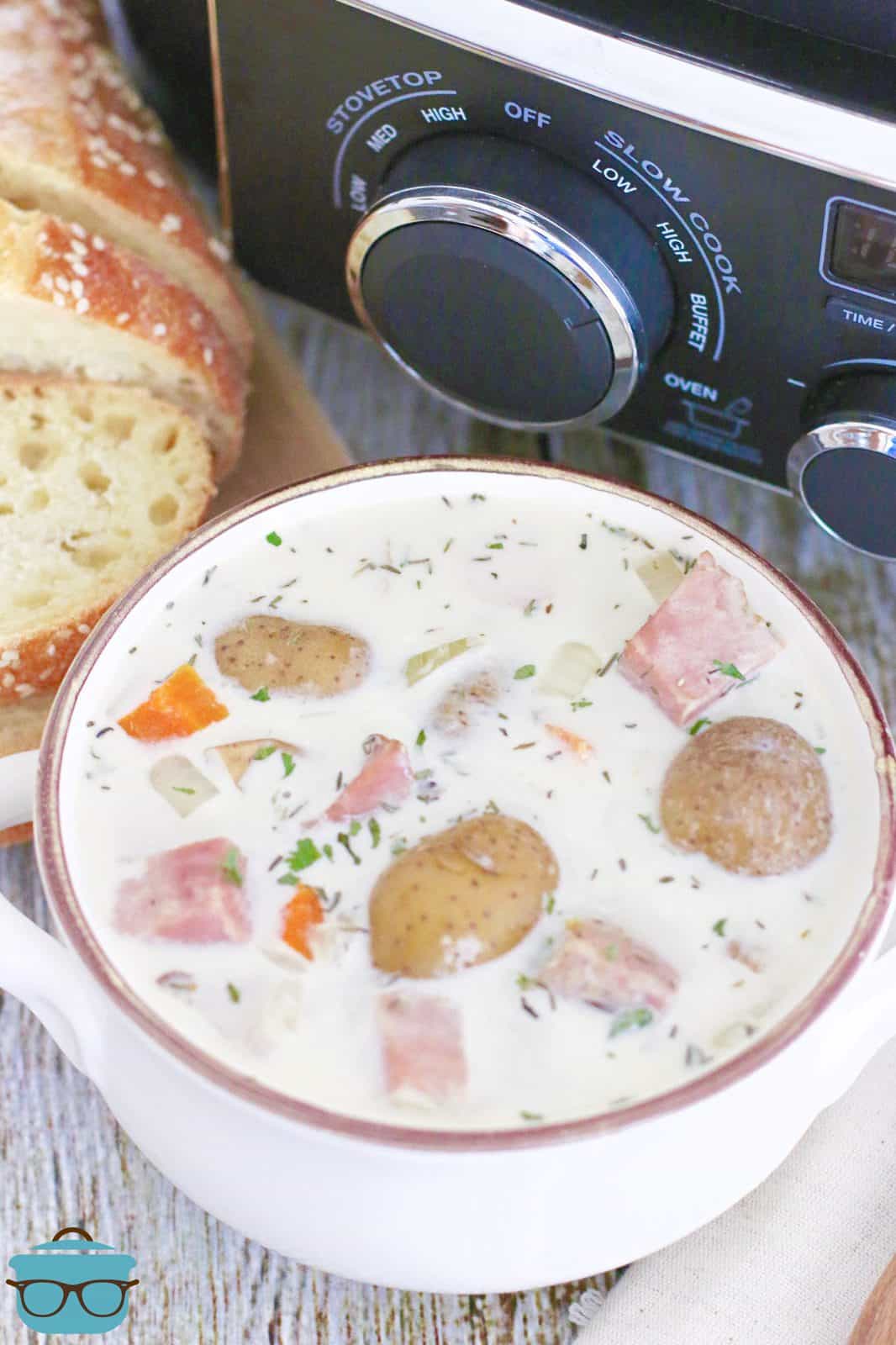 a white bowl of cream potato and ham soup in front of a black slow cooker.