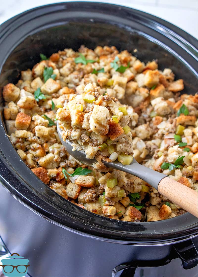 Crock Pot Stuffing recipe shown in an oval slow cooker, spoon scooping some stuffing out of the slow cooker.
