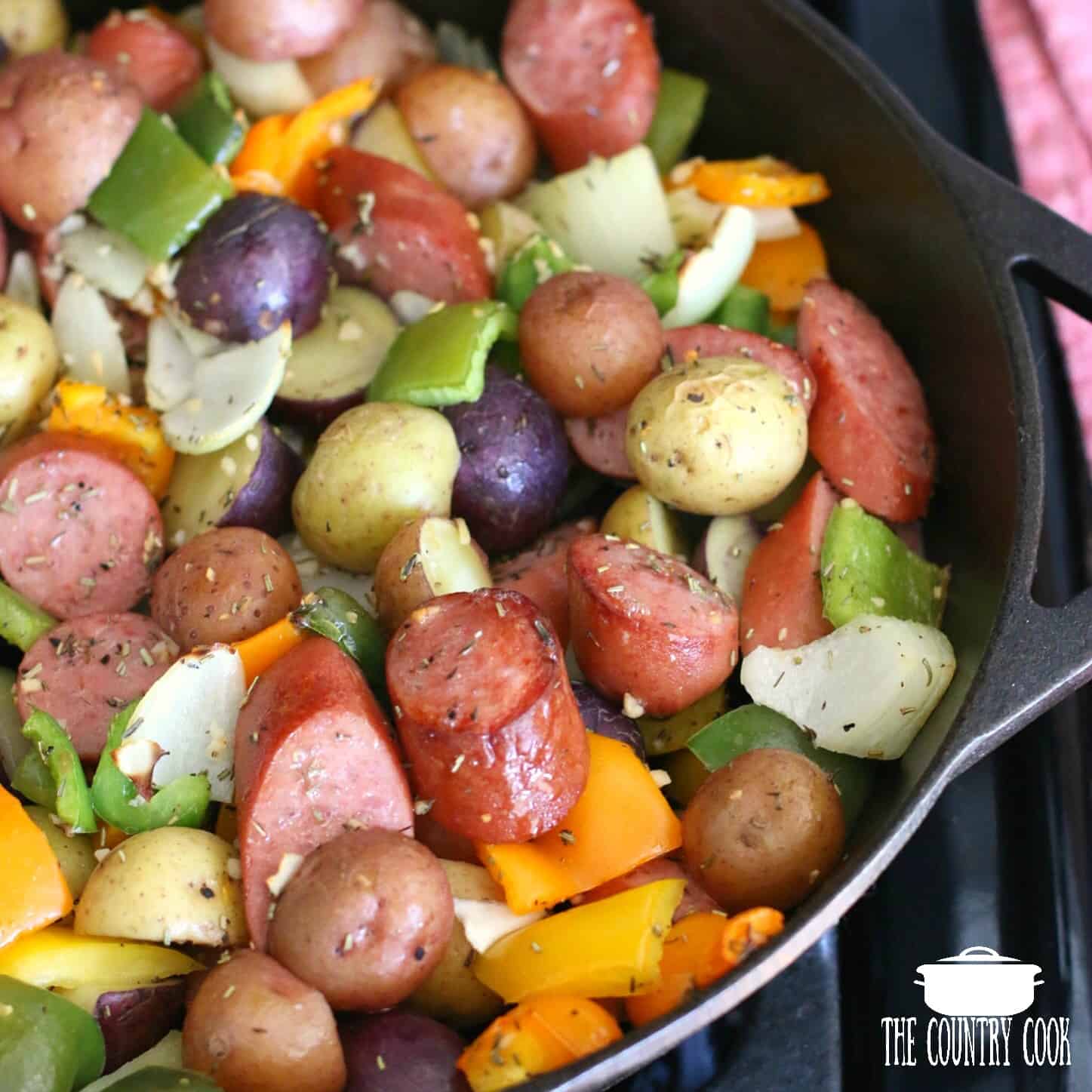 sliced kielbasa, baby potatoes, peppers and onions shown close up in a cast iron skillet.