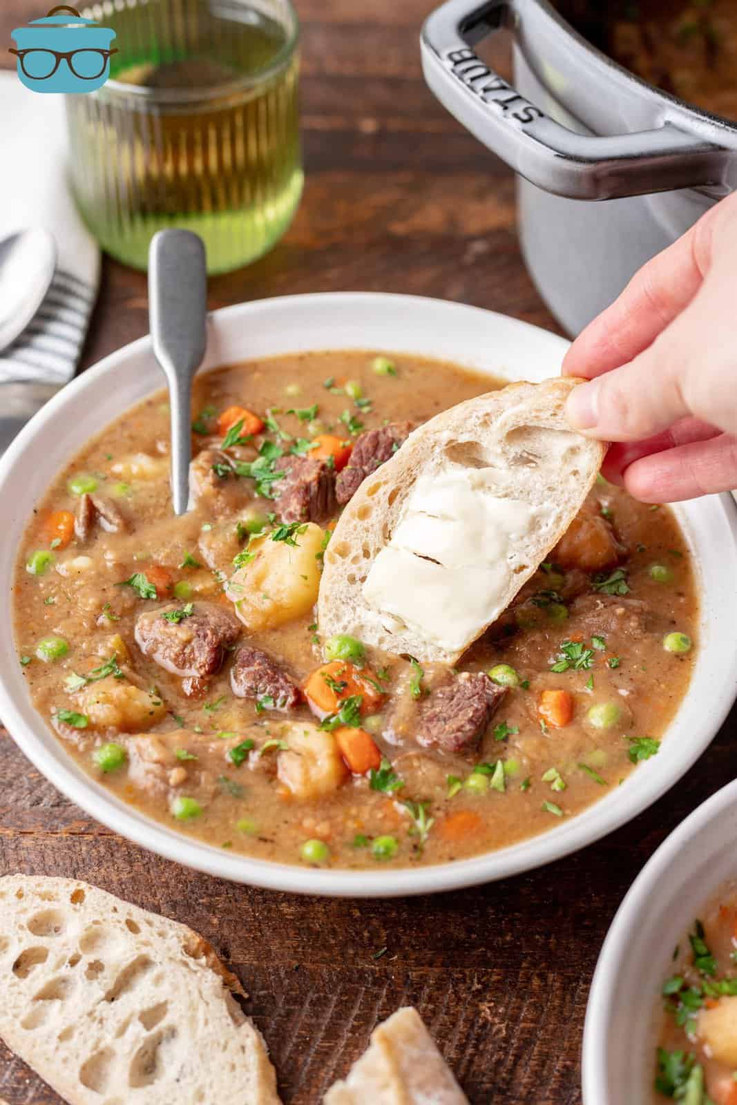 a white bowl of beef stew with a hand to the side of the bowl holding up a piece of a slice of buttered French bread. 