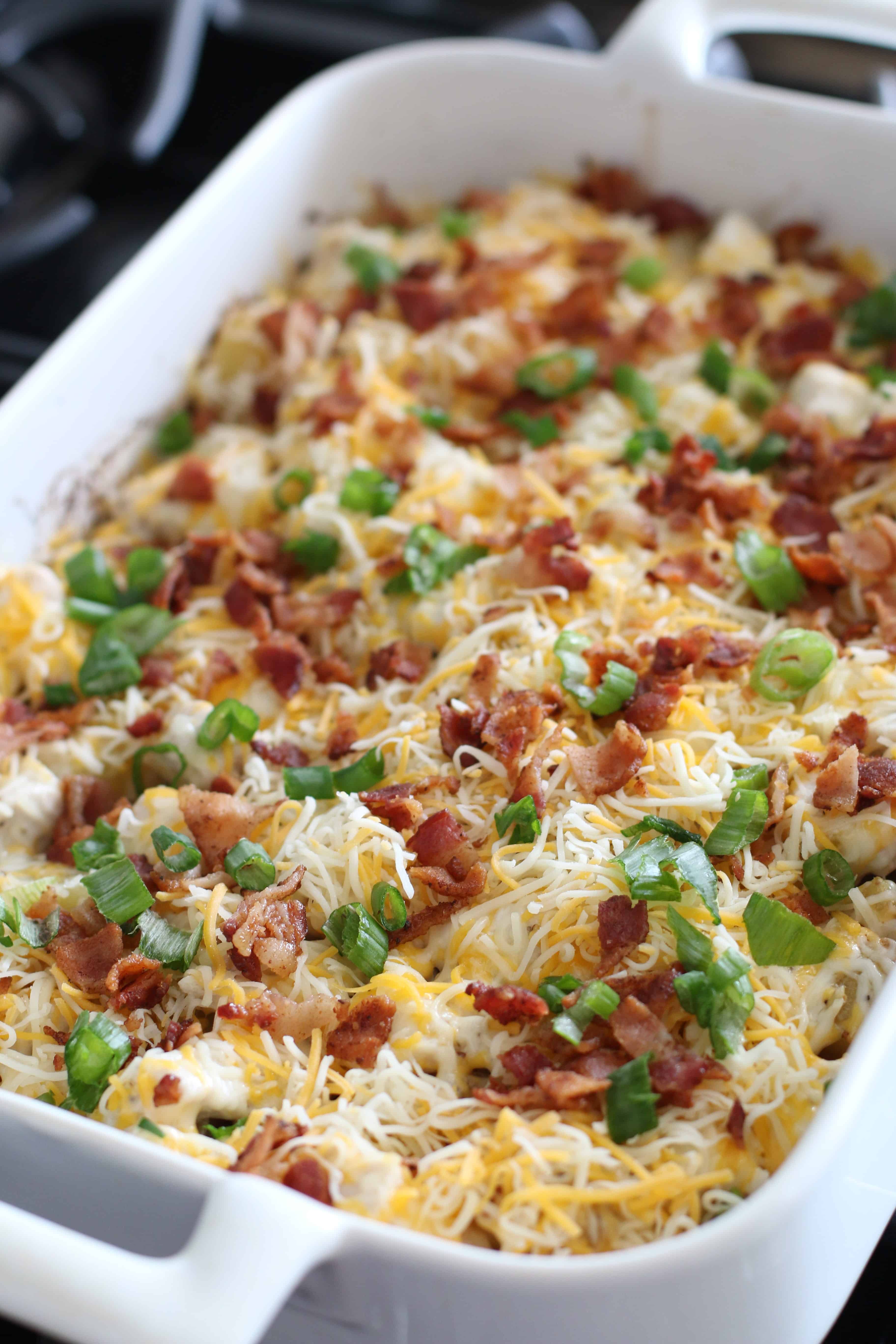 shredded cheese, bacon and green onion with diced potatoes and chicken in a white baking dish. 