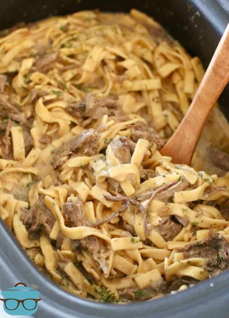 roast beef and cooked egg noodles shown in an oval slow cooker with a wooden spoon
