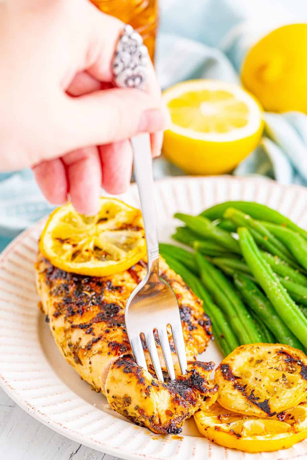 hand holding a fork to cut a slice of grilled lemon garlic chicken on a white plate with cooked green beans on the side.