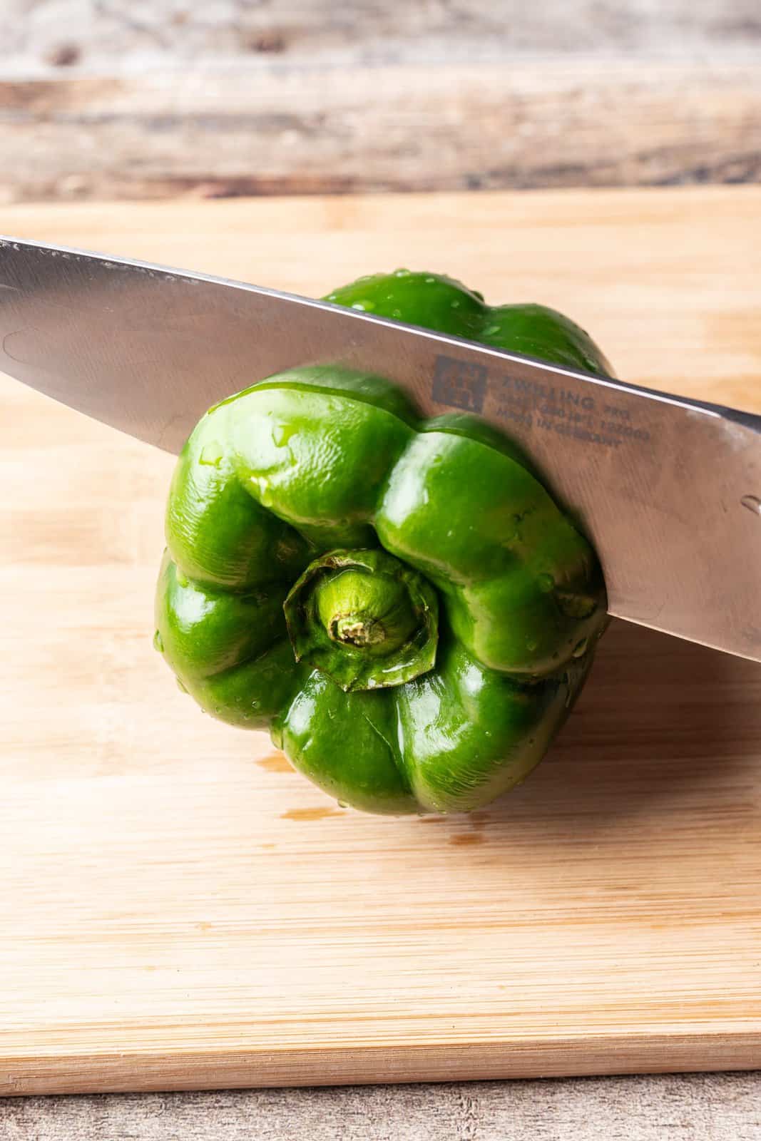 cutting off the top of a green pepper with a knife on a wooden cutting board. 