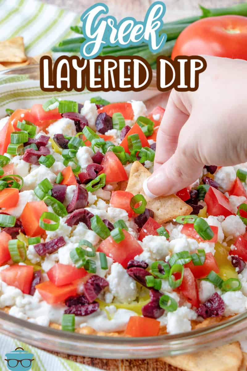 a hand dipping in a pita chip into the pan of prepared Greek Layered Dip.
