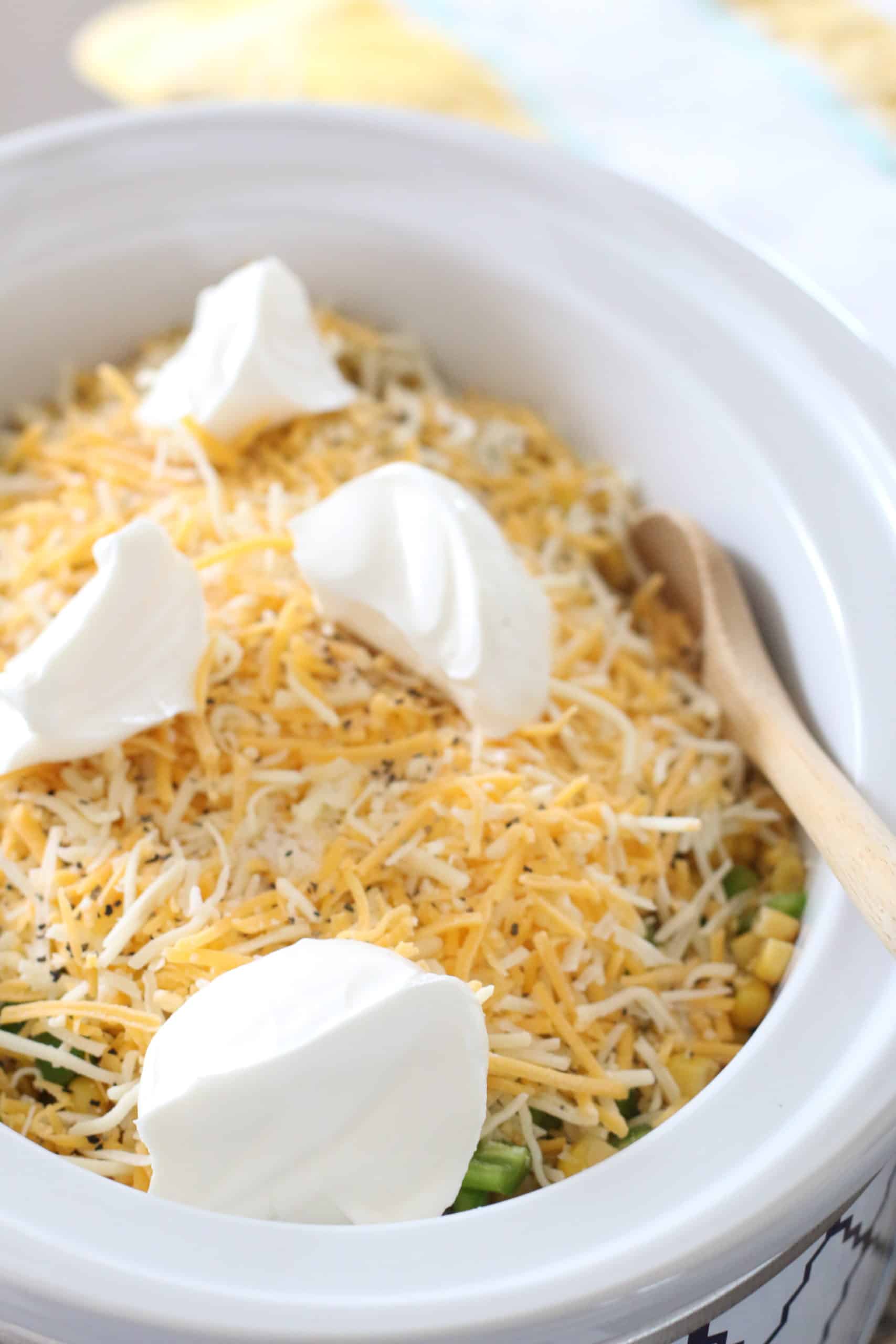 four large dollops of sour cream on top of shredded cheese in slow cooker with wooden spoon on the side.