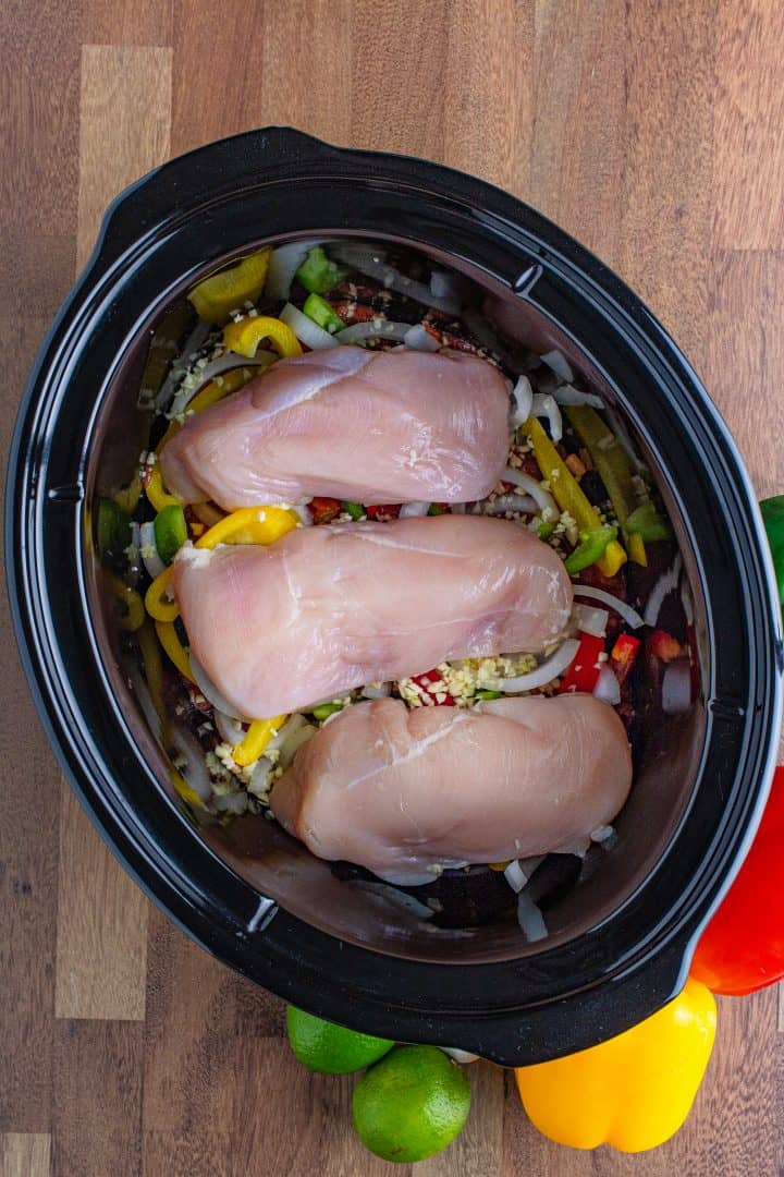 three fresh chicken breasts laid on top of sliced onions and peppers in an oval slow cooker. 