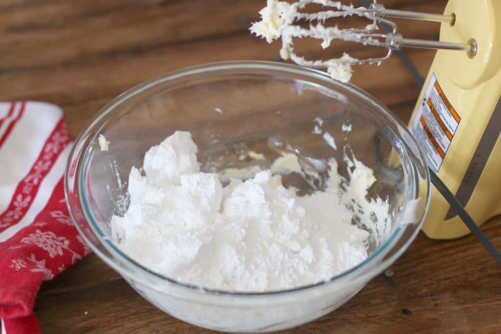 confectioners sugar added into softened butter and cream cheese in a clear Pyrex bowl.