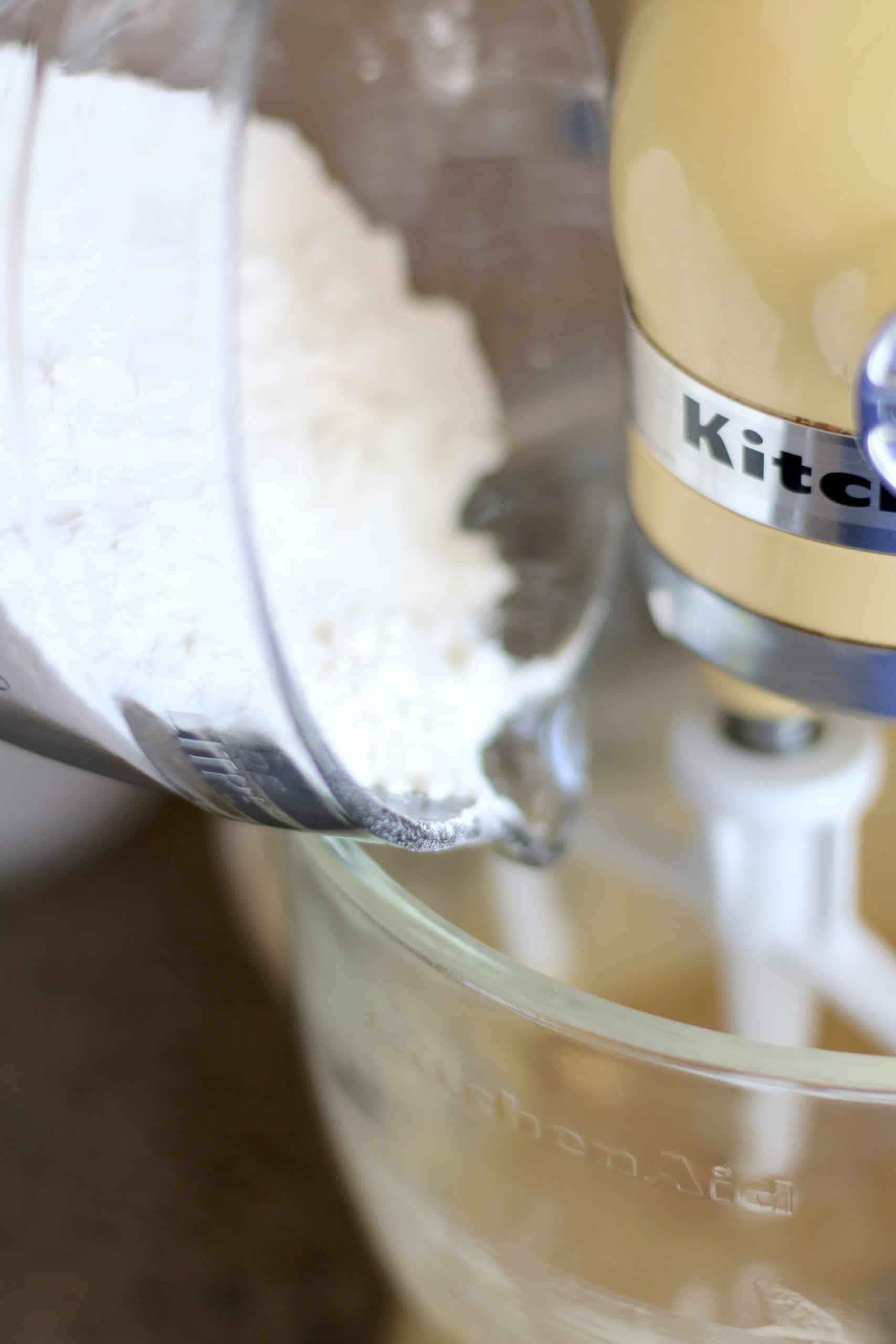 pouring in flour mixture into a clear mixing bowl of a yellow stand mixer.