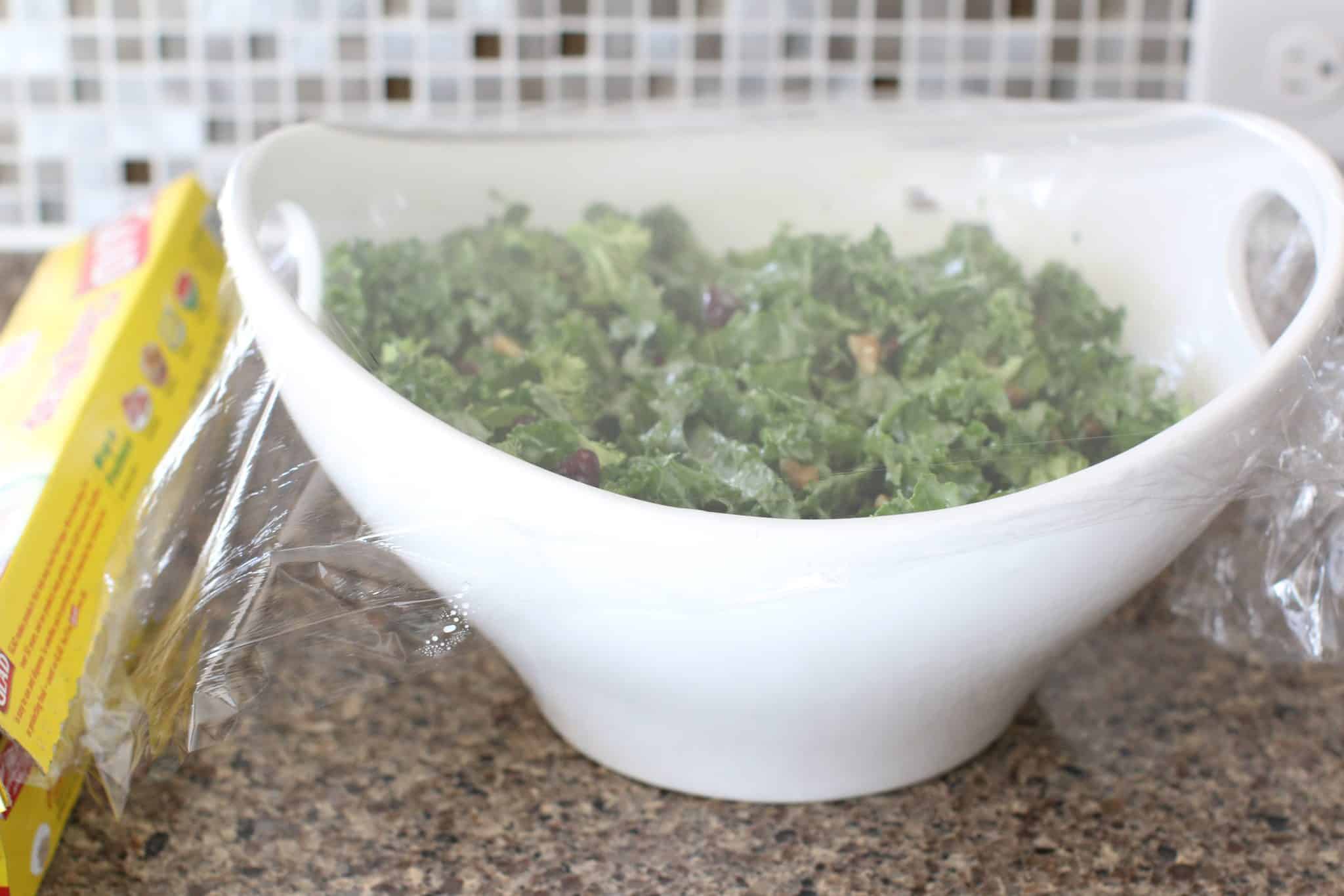 kale salad shown in a white bowl with handles and covered in plastic wrap. 