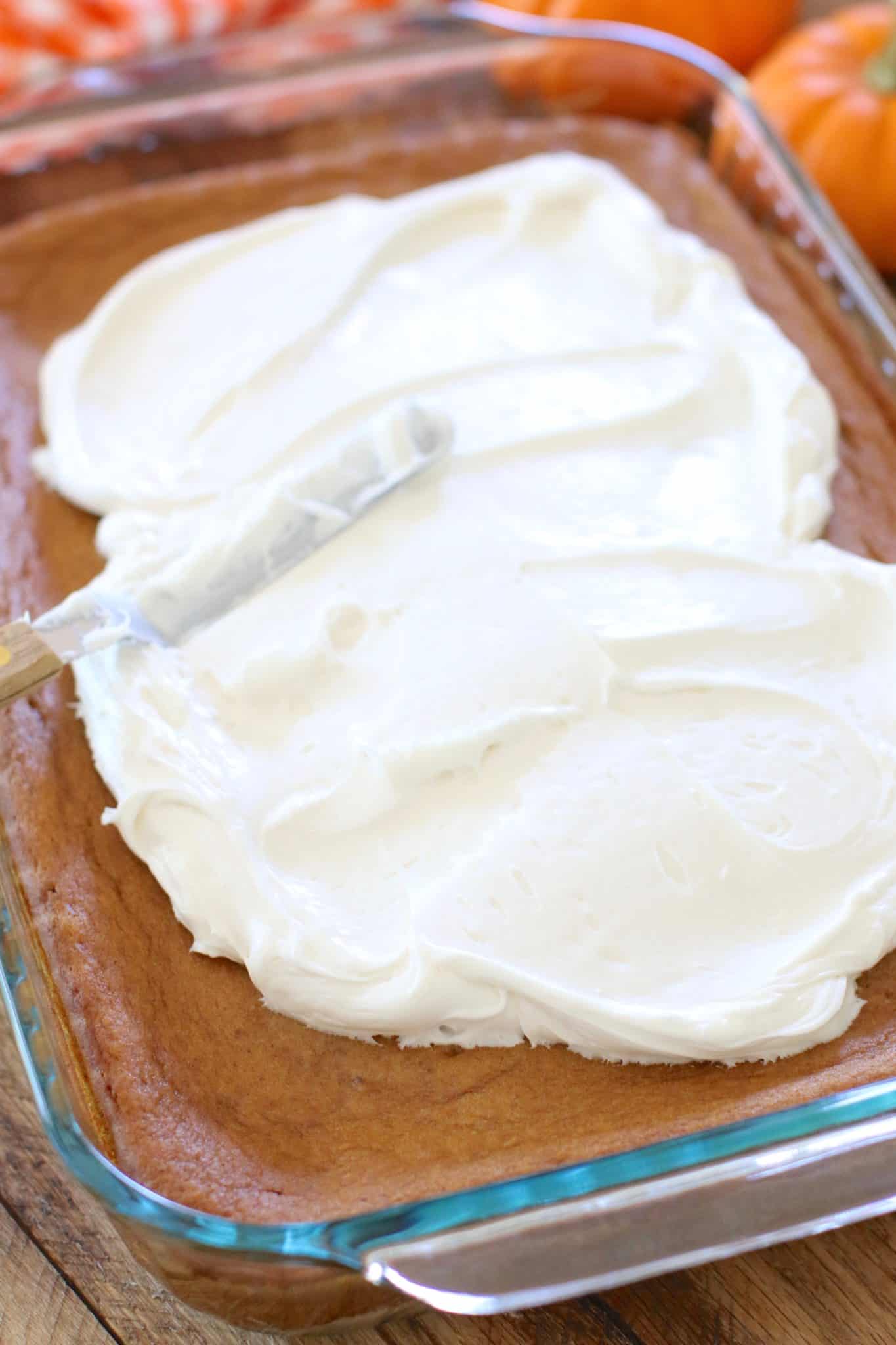 spreading white premade frosting on cooled pumpkin pie cake with an offset spatula. 