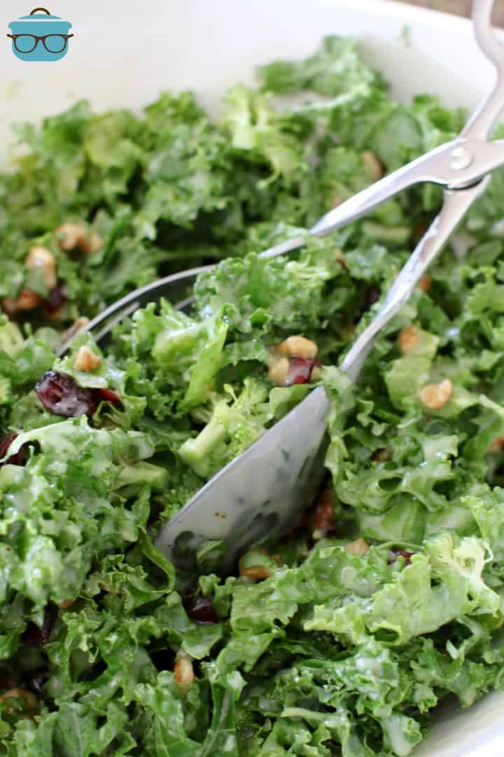 a pair of silver salad tongs shown in a white bowl filled with kale salad. 