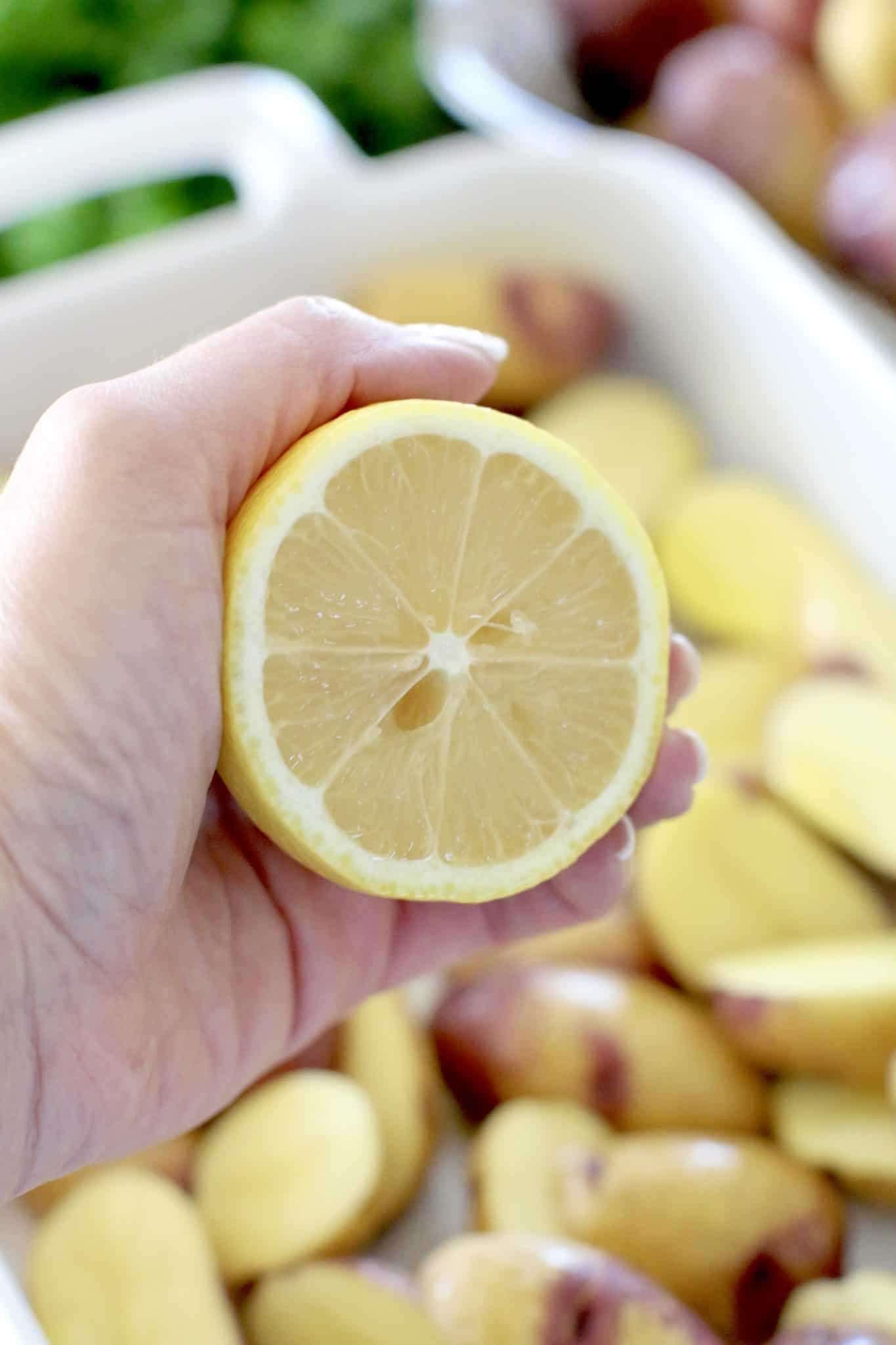 a hand squeezing lemon juice out of half a lemon over sliced potatoes in baking dish.