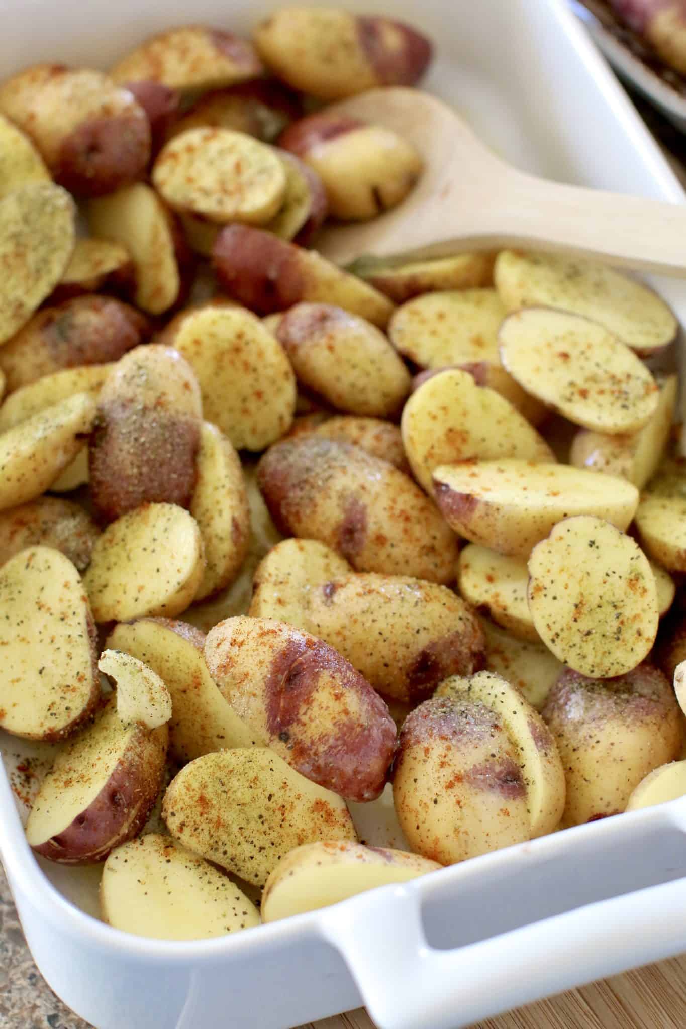 Greek seasoning shown sprinkled over sliced potatoes in a white baking dish and a wooden spoon on the side. 