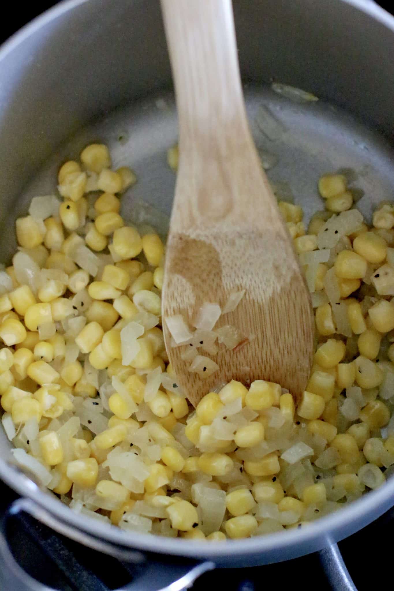 corn kernels and chopped onion in a pot being stirred by a wooden spoon.