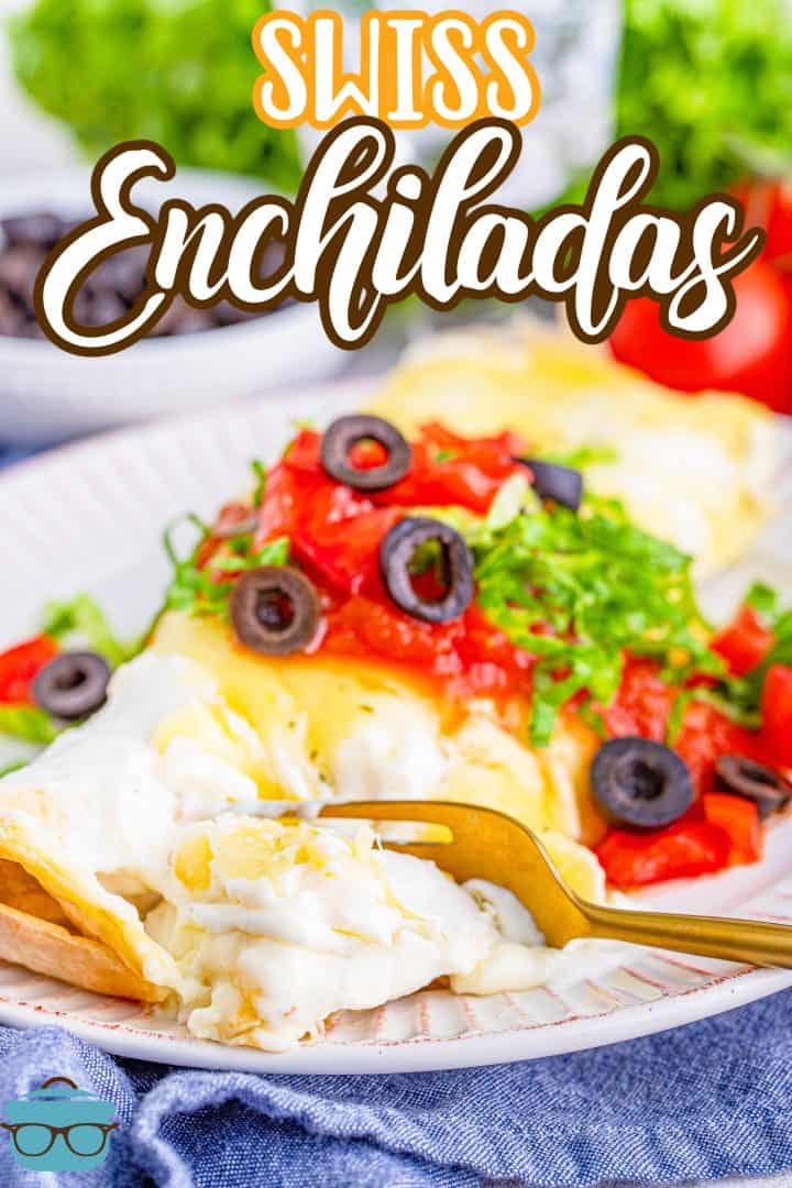 two Swiss enchiladas on a white plate and topped with salsa, olives and sliced lettuce with a fork starting to scoop up a bite. 