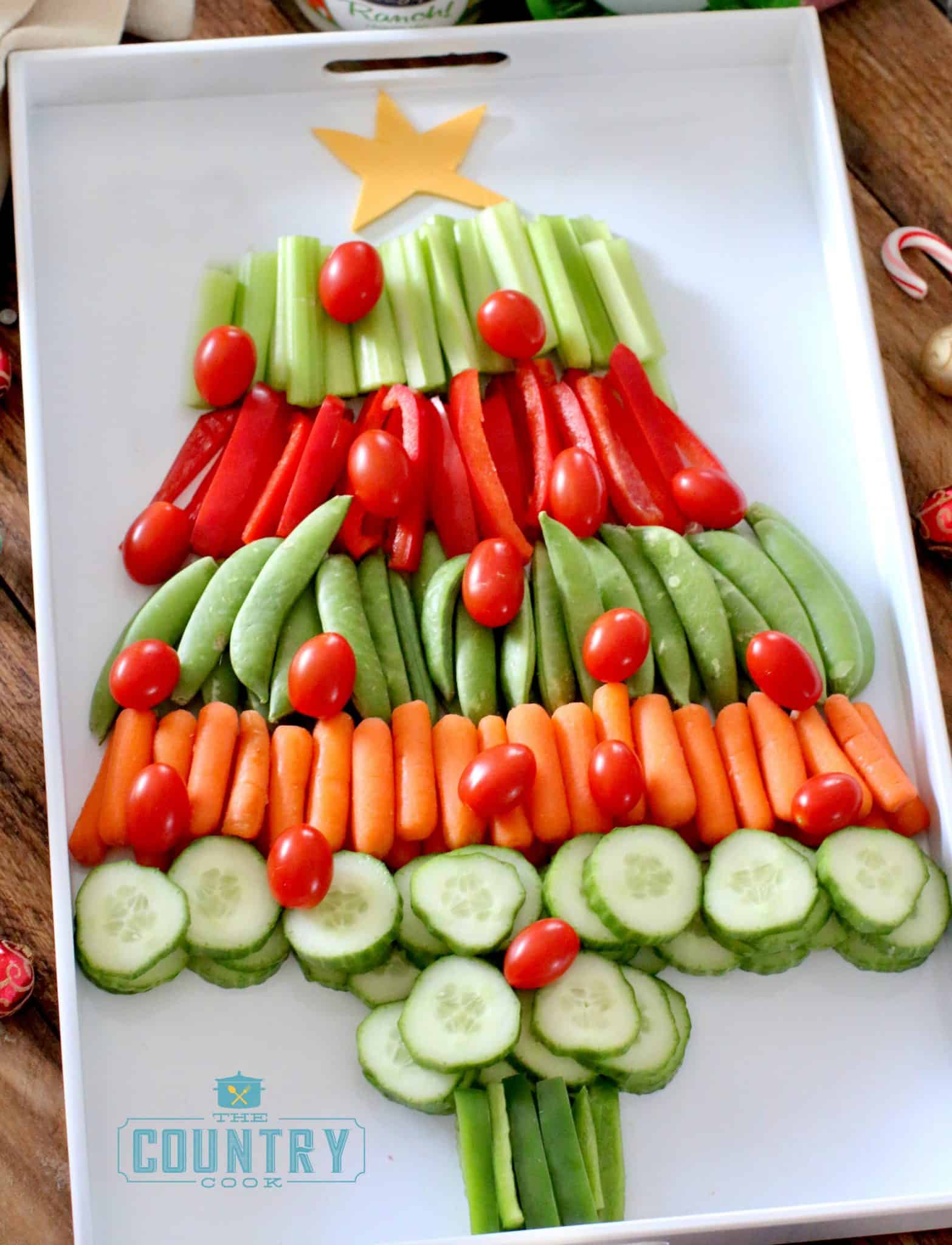 layers of cucumbers, carrots, snap peas, red peppers and celery to make the shape of a Christmas tree on a white tray.