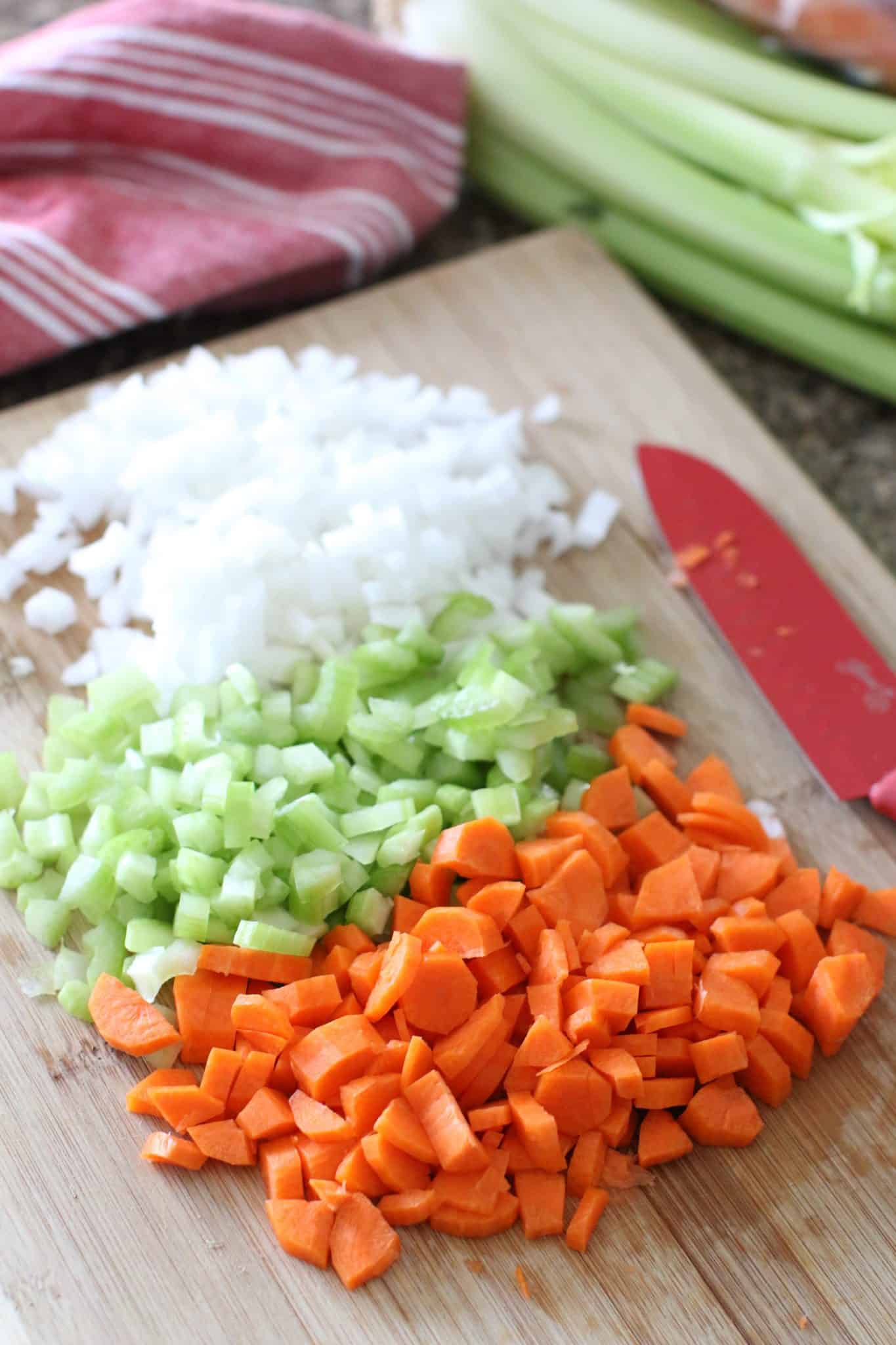 diced onion, celery, carrots on a wooden cutting board with a red knife.