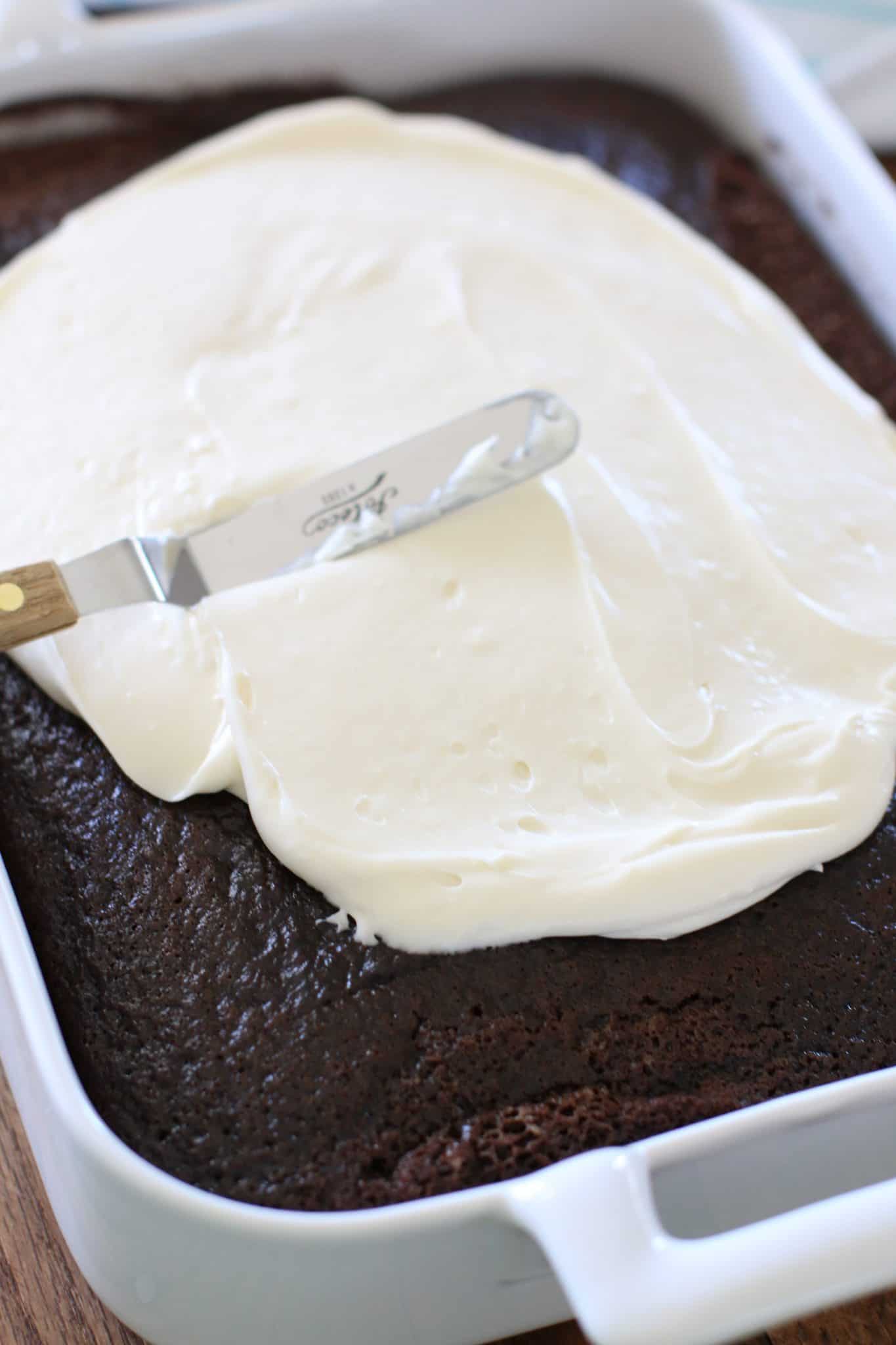 an offset spatula spreading homemade frosting onto a baked chocolate cake.