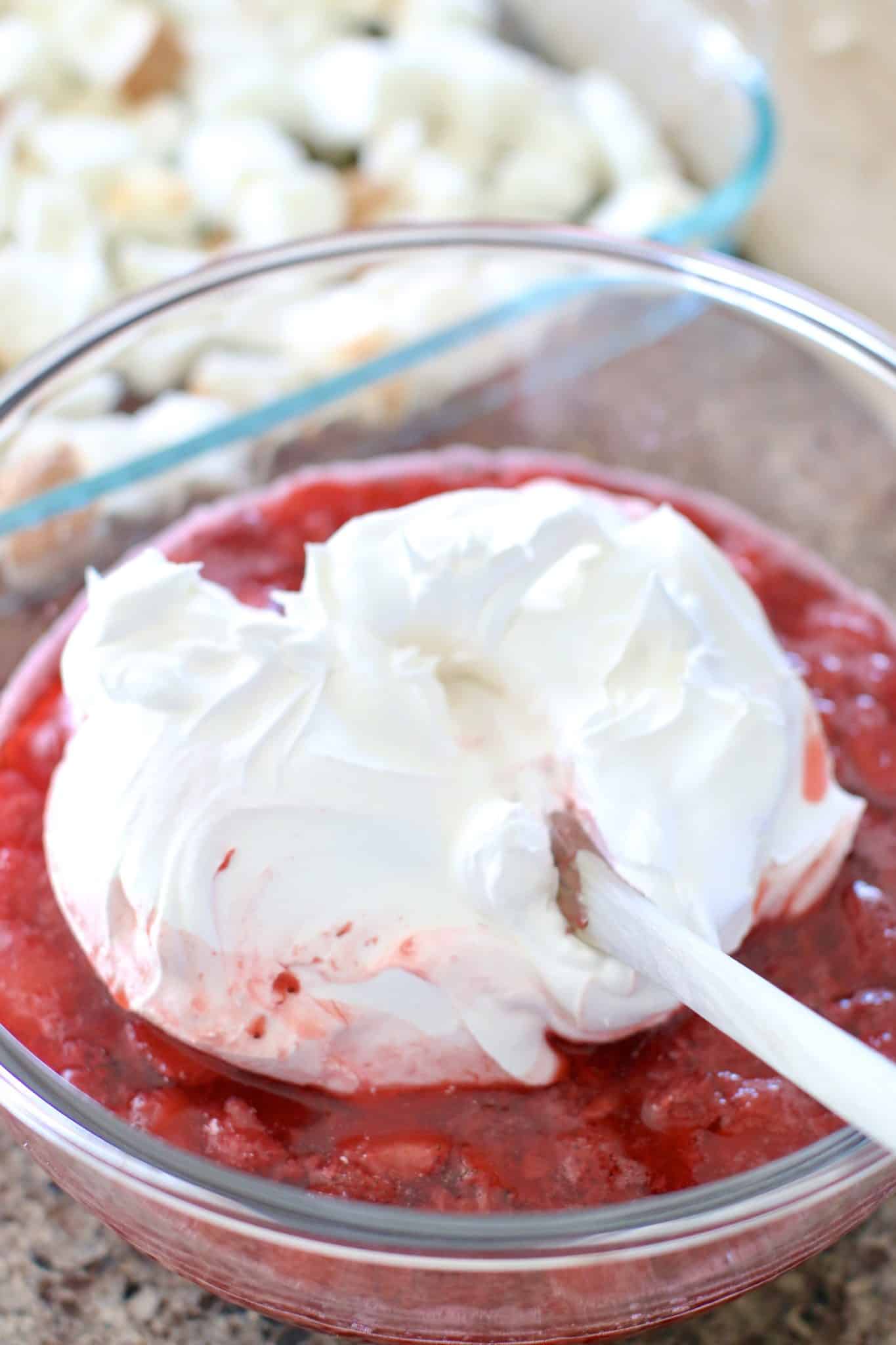 cool whip whipped topping added to frozen strawberries and strawberry gelatin mixture in a glass mixing bowl with a wooden spoon.