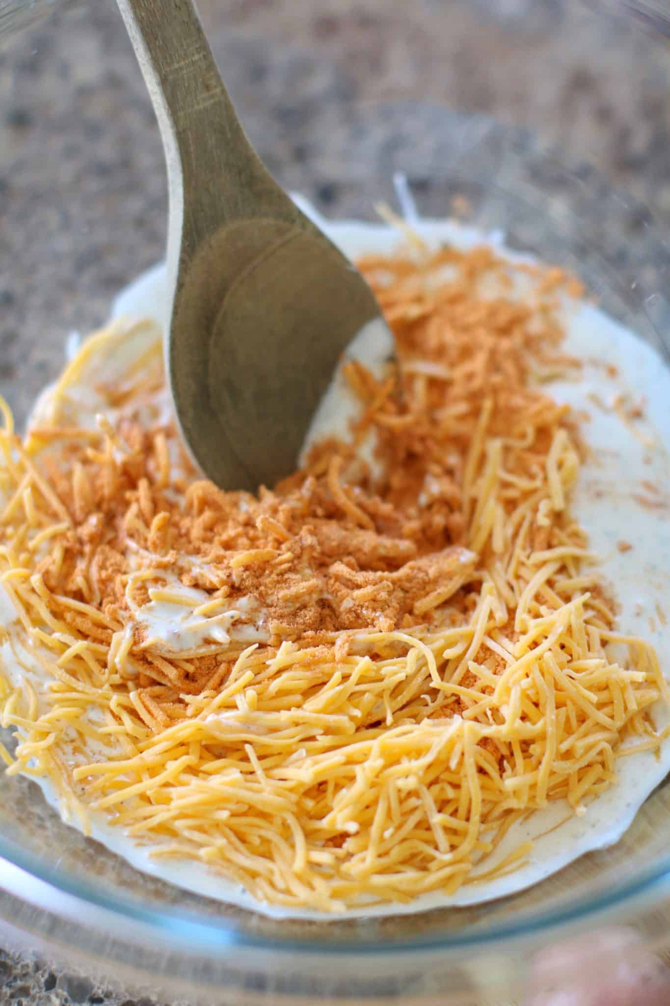 a wooden spoon in a clear bowl with taco seasoning, ranch dressing and shredded cheese. 