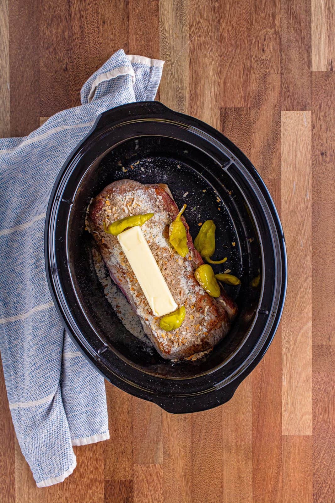 stick of butter placed on a pork roast with peperroncini peppers in an oval slow cooker.