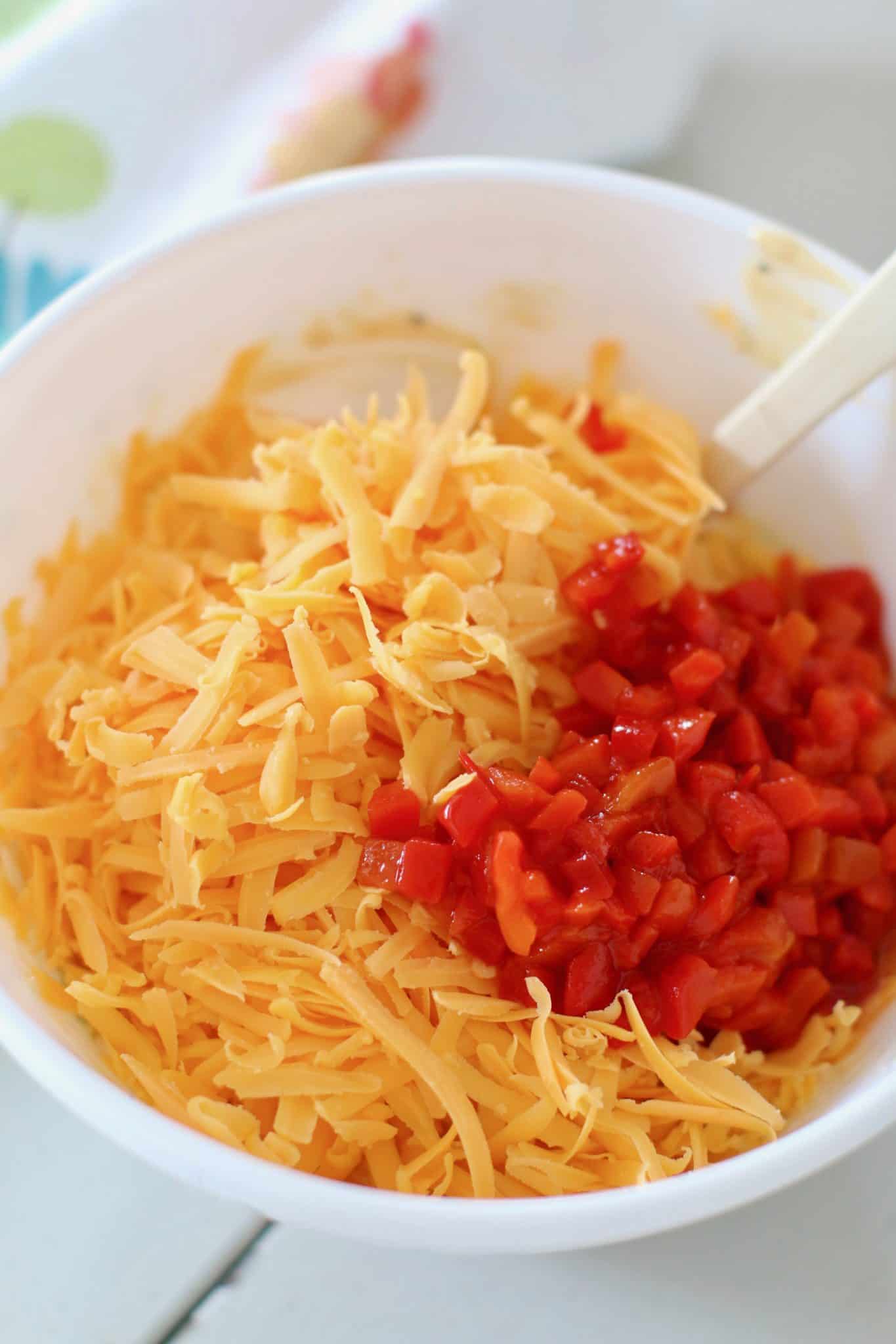 shredded cheddar cheese and diced pimentos added to cream cheese mixture in a white bowl.