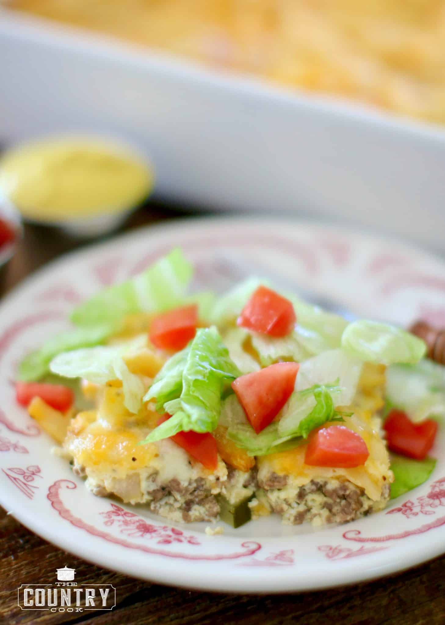 A slice of cheeseburger and fries casserole served on a plate and topped with shredded lettuce and diced tomatoes.