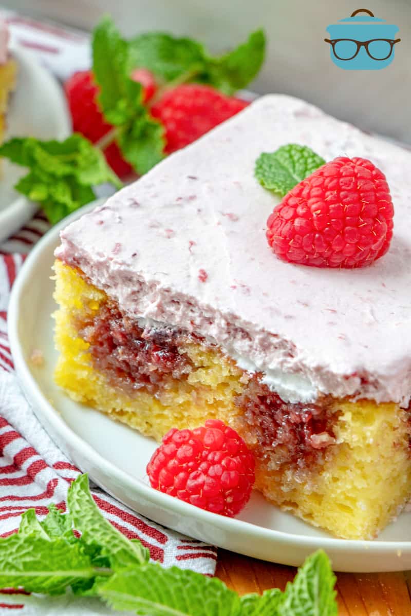 Lemon Raspberry Poke Cake - slice shown on a white plate, slice topped with a fresh raspberry with a mint leaf.