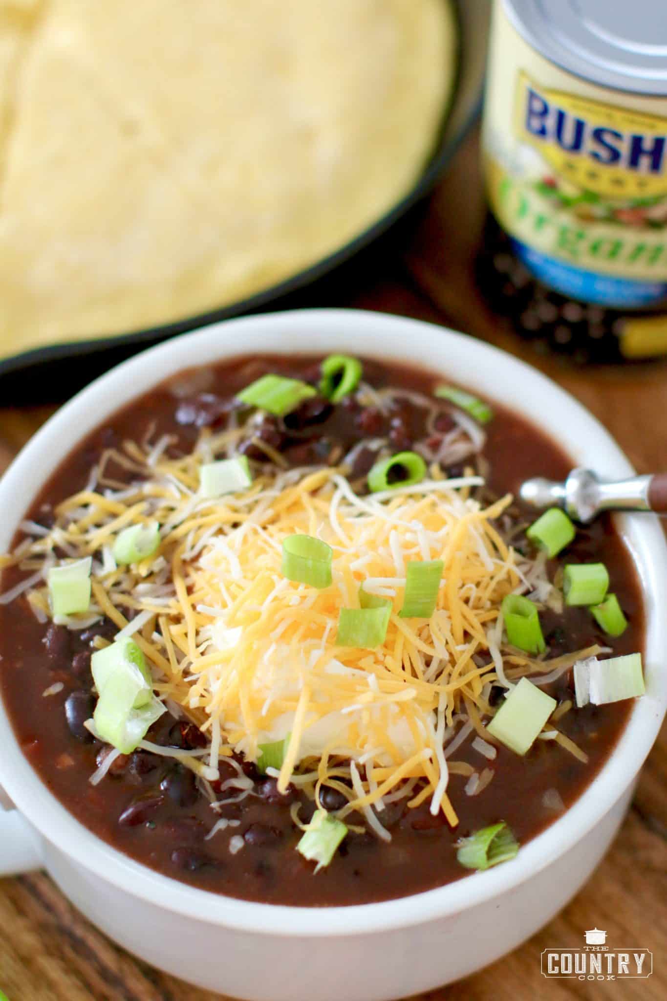 Black Bean Chili in a bowl topped with shredded cheese and green onions with a pan of cornbread in the background.