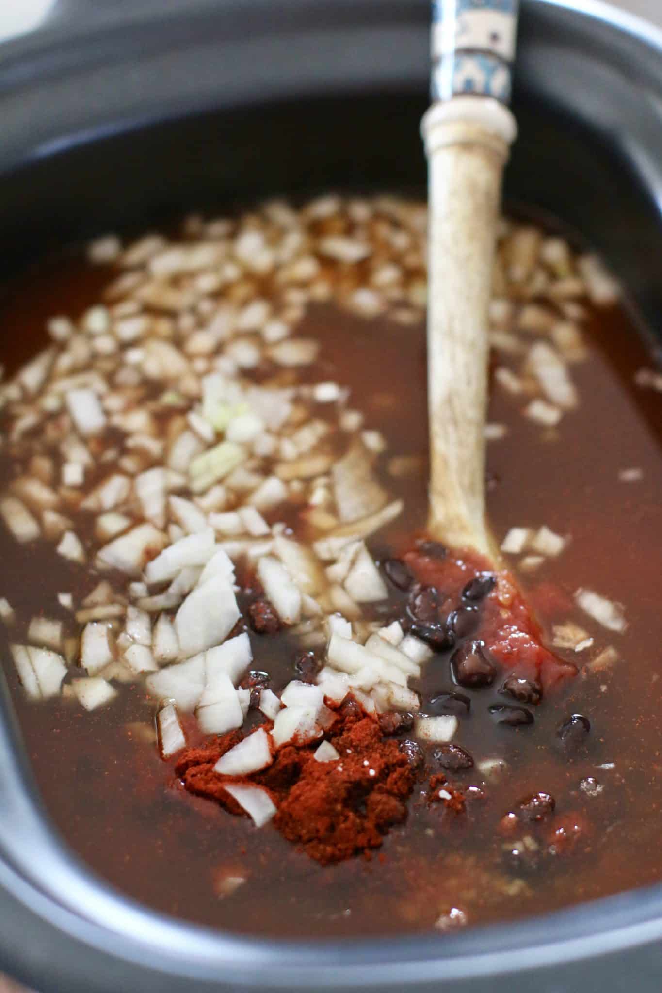 ingredients for black bean chili in crock pot being stirred by a wooden spoon.