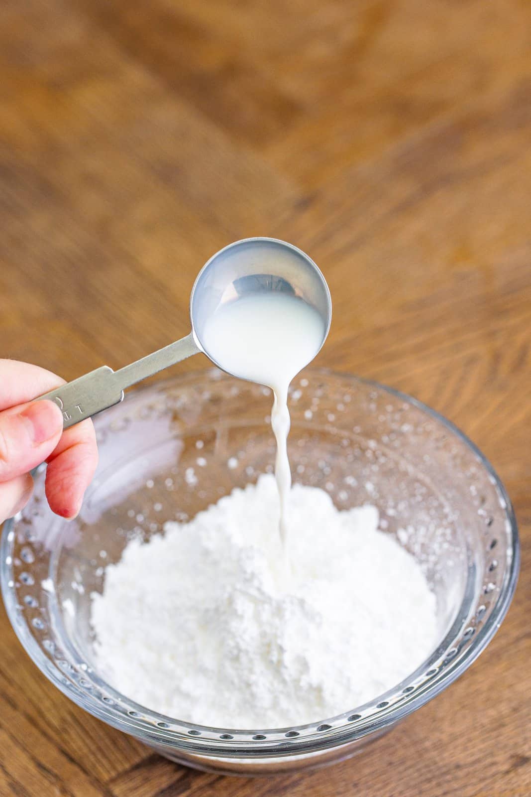 a measuring spoon pouring milk into the powdered sugar in a bowl.