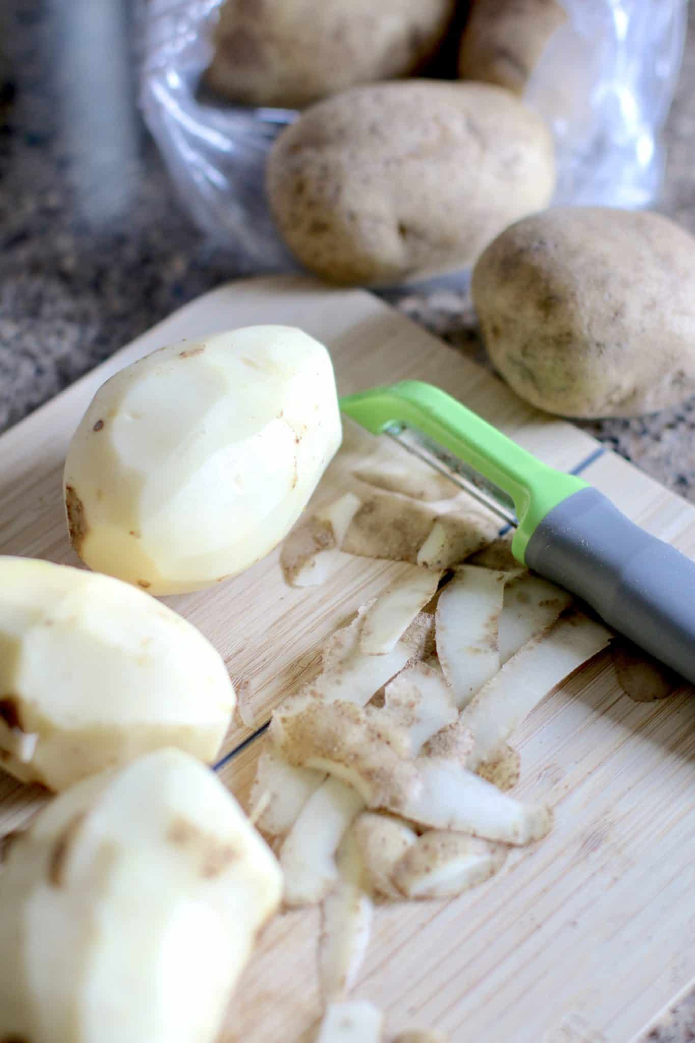 peeled potatoes shown on a wooden cutting board with a potato peeler.