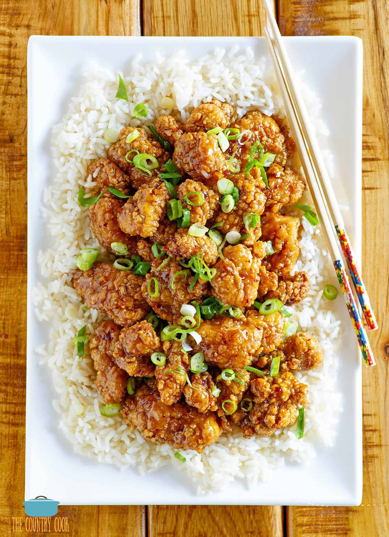 Honey Garlic Glazed Asian Popcorn Chicken on top of white rice shown on a large white rectangle plate with chopsticks on the side.