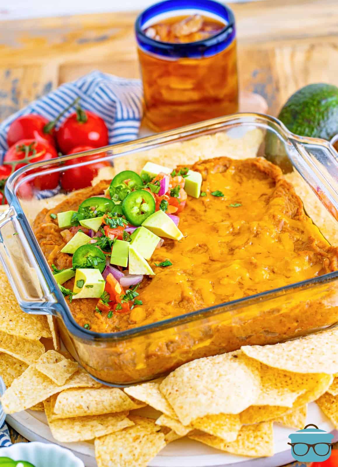 fully baked refried bean dip with toppings shown with tortilla chips and a glass of iced tea in the background. 
