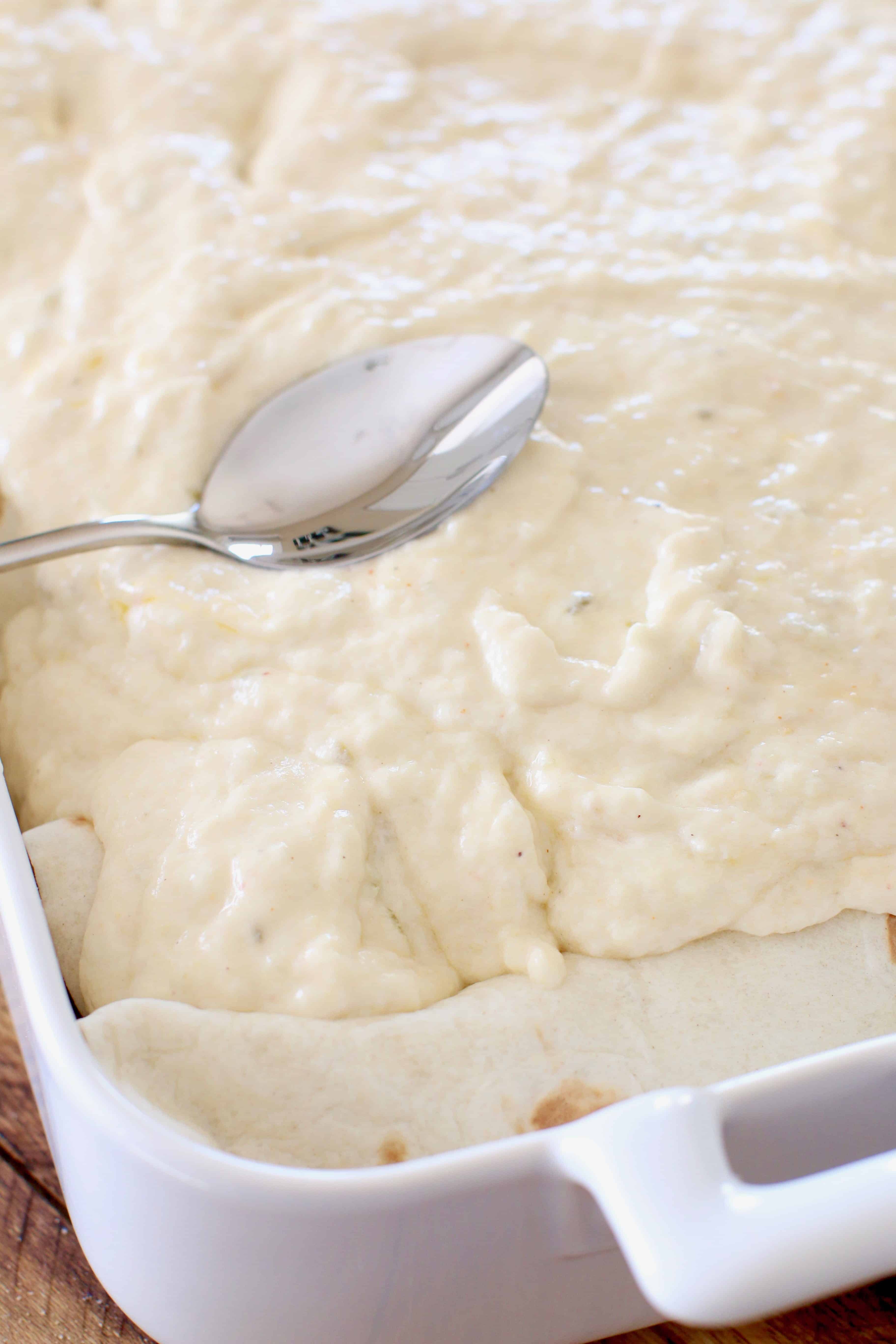 creamy cheese sauce on top of shrimp enchiladas being spread out with the back of a silver spoon.