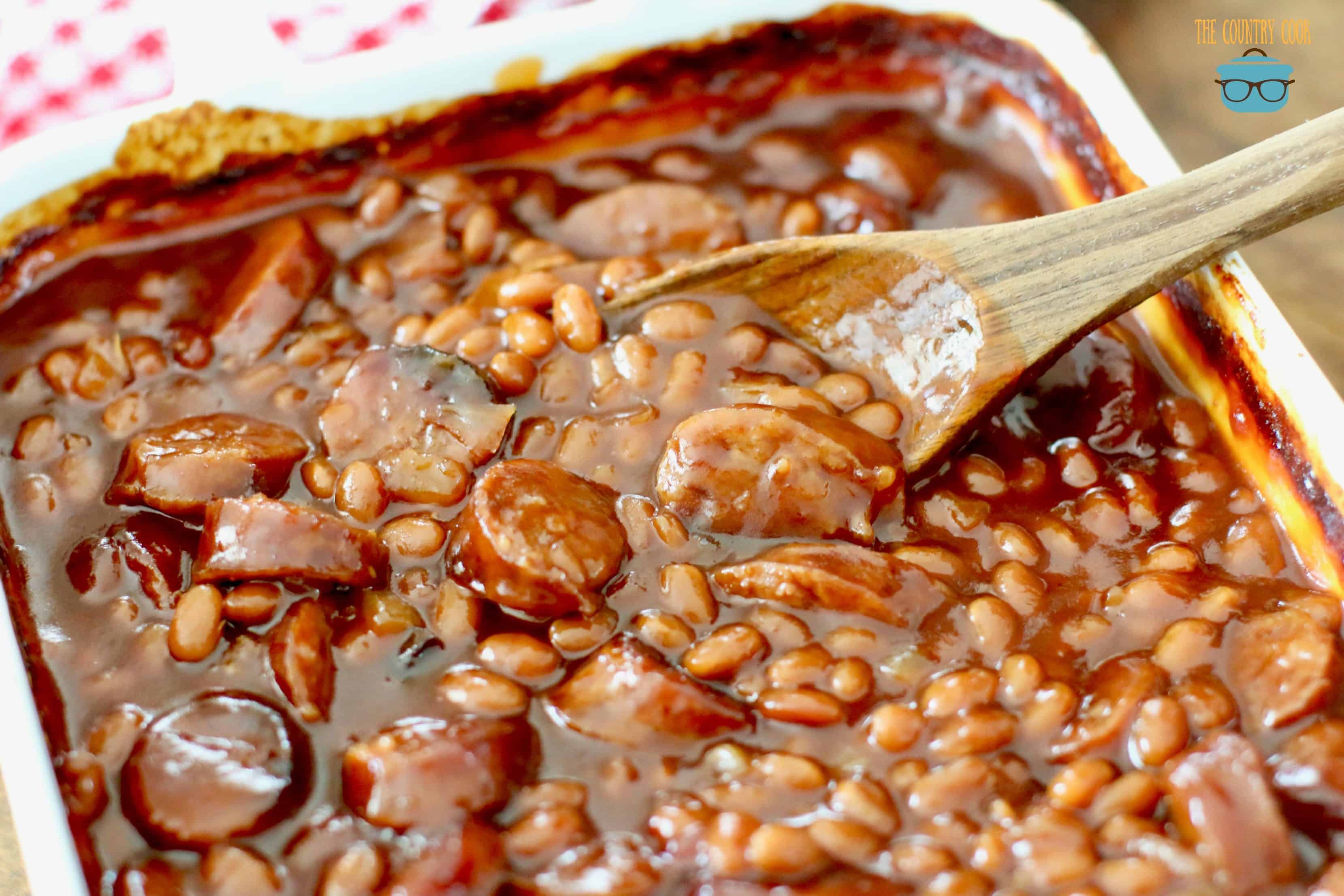baked beans in a white baking dish with a wooden spoon inserted. 