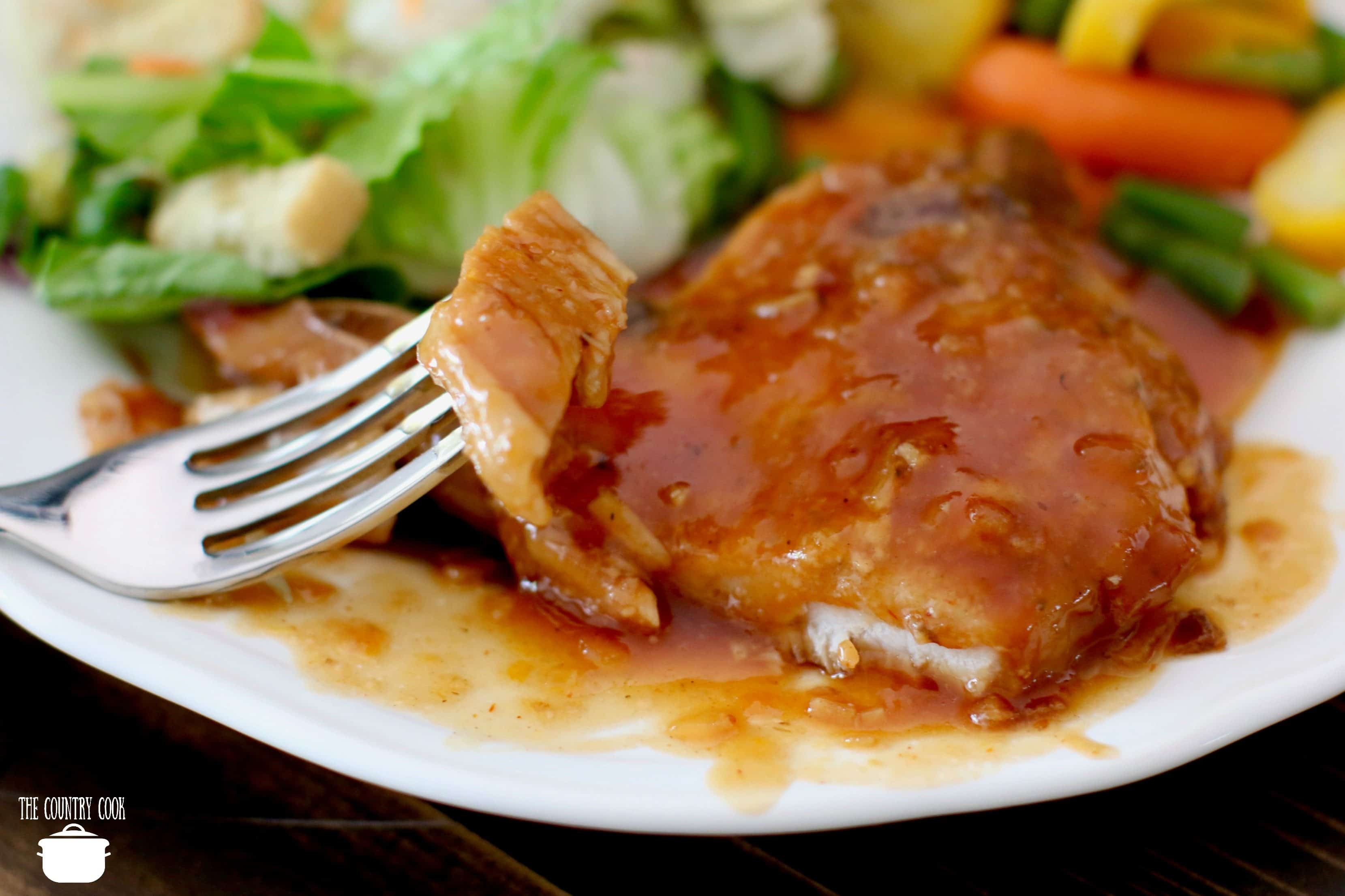 Fork shown holding up a slice of BBQ pork chops on a white plate with a salad and vegetables in the background.