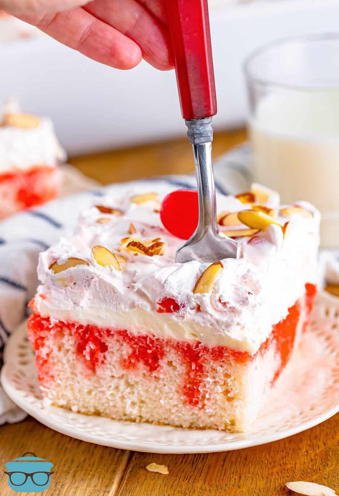 a fork shown being inserted by a hand into a slice of cherry almond poke cake that is on a white plate. 