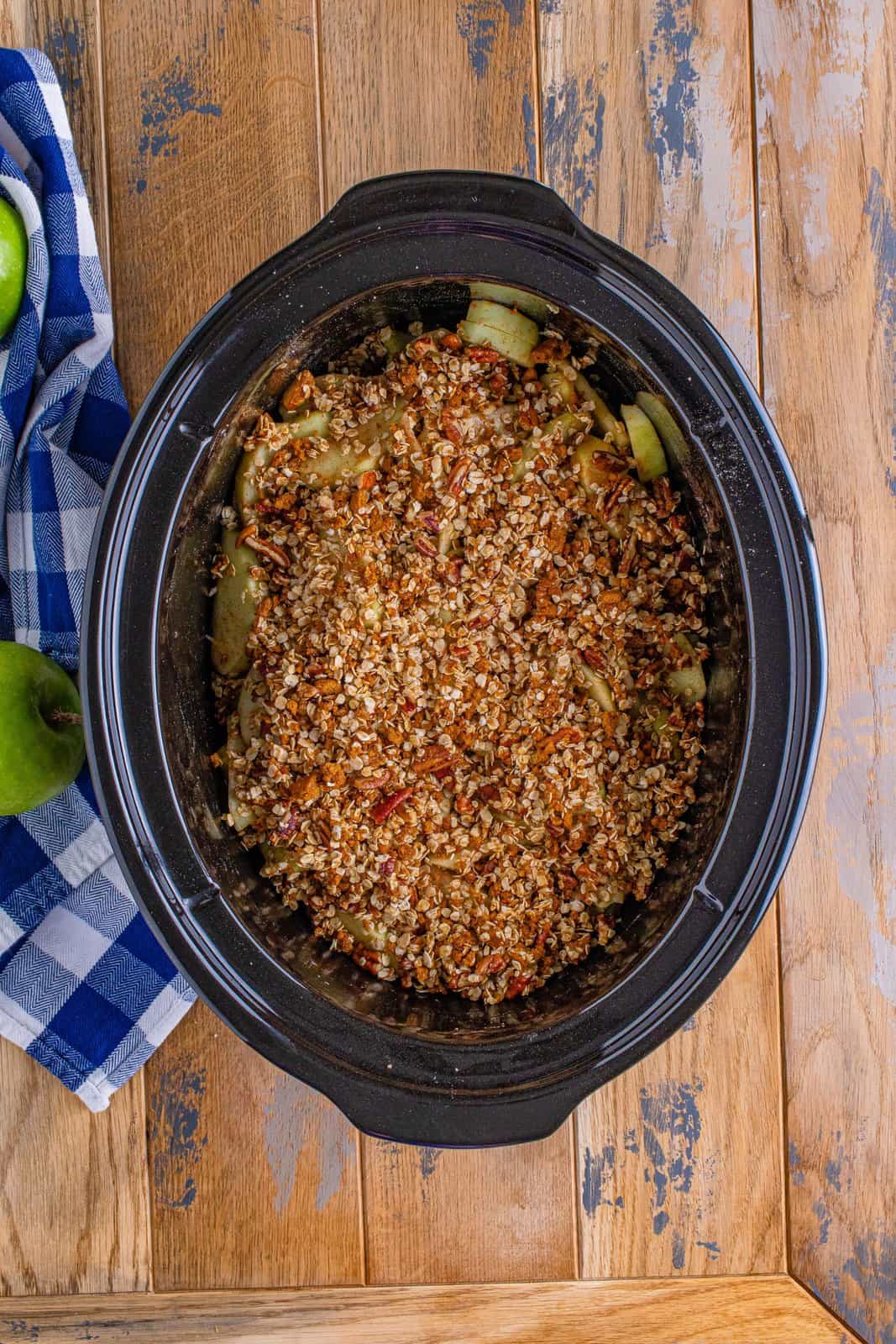 oatmeal topping sprinkled over sliced apple mixture in an oval slow cooker.