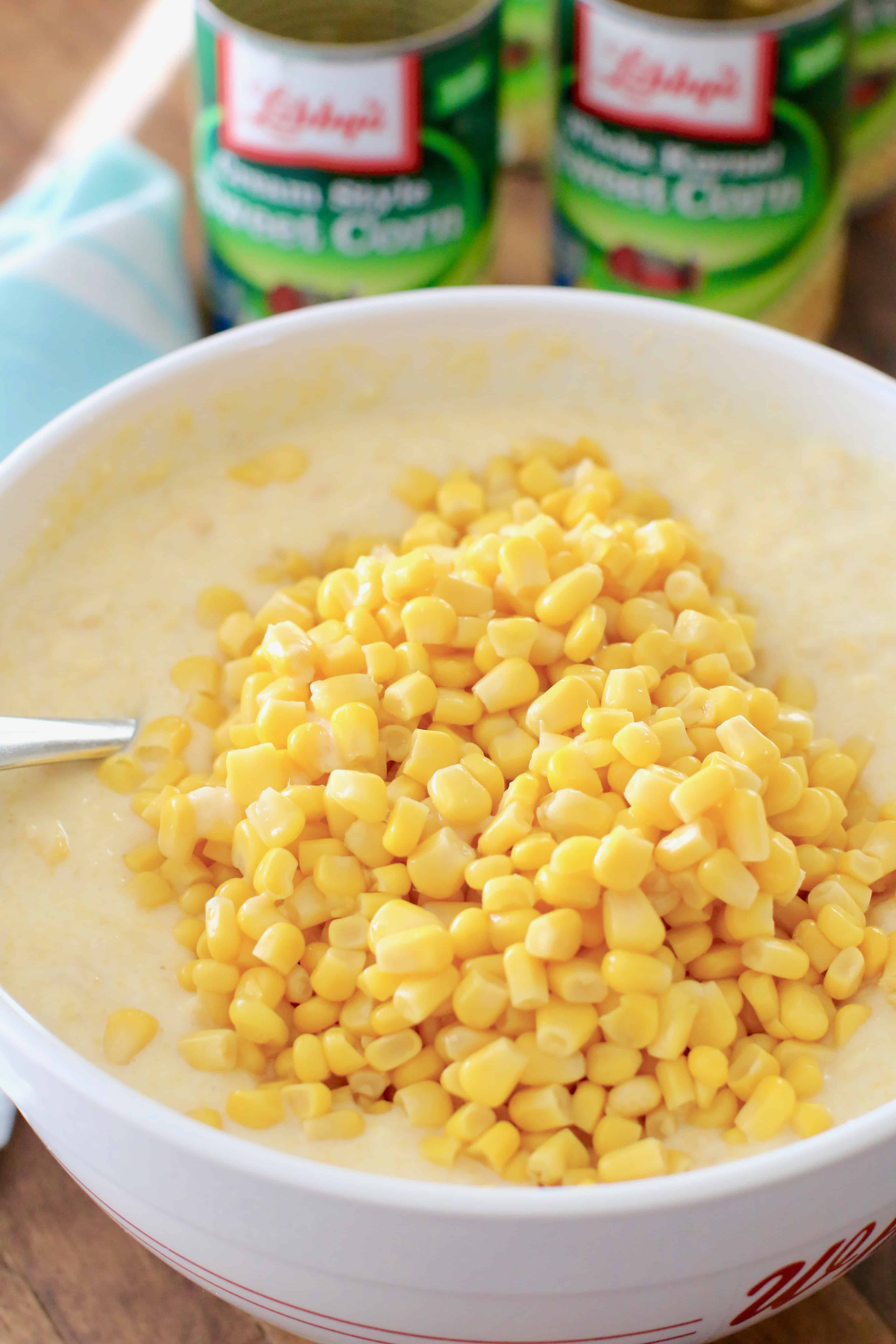 sweet corn casserole batter with canned corn down in a white mixing bowl with cans of corn in the background.