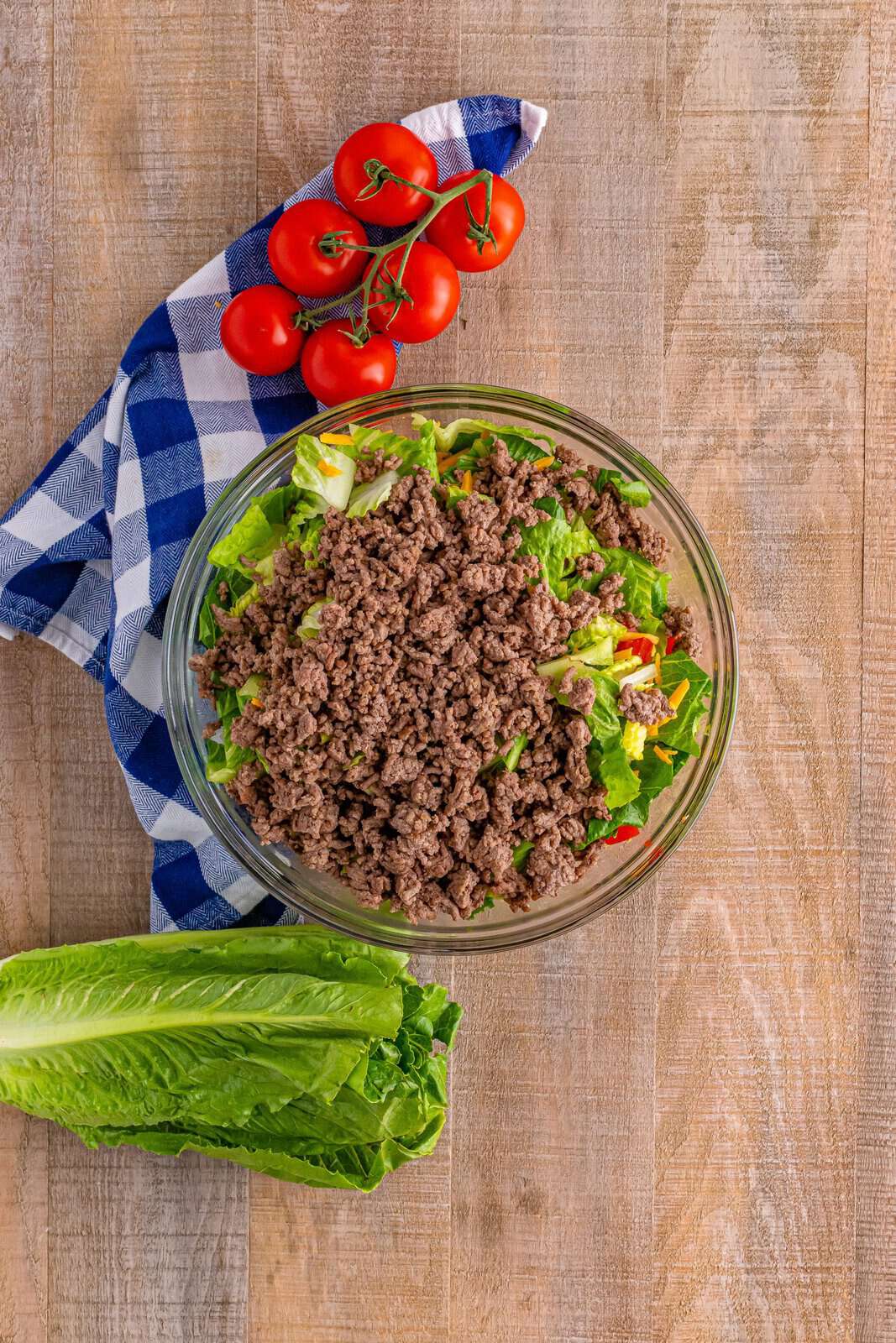 cooked ground beef added to salad ingredients in a large glass bowl. 