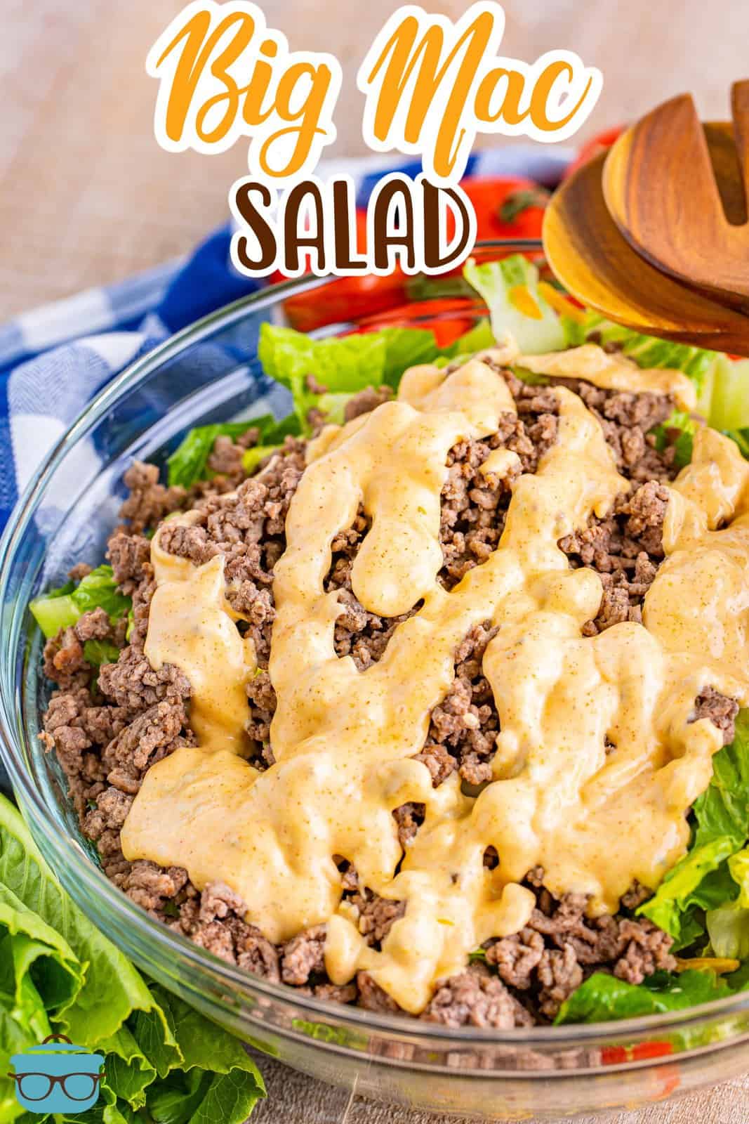 a glass bowl filled with salad ingredients topped with ground beef and drizzled with Big Mac Sauce with salad tongs in the background. 